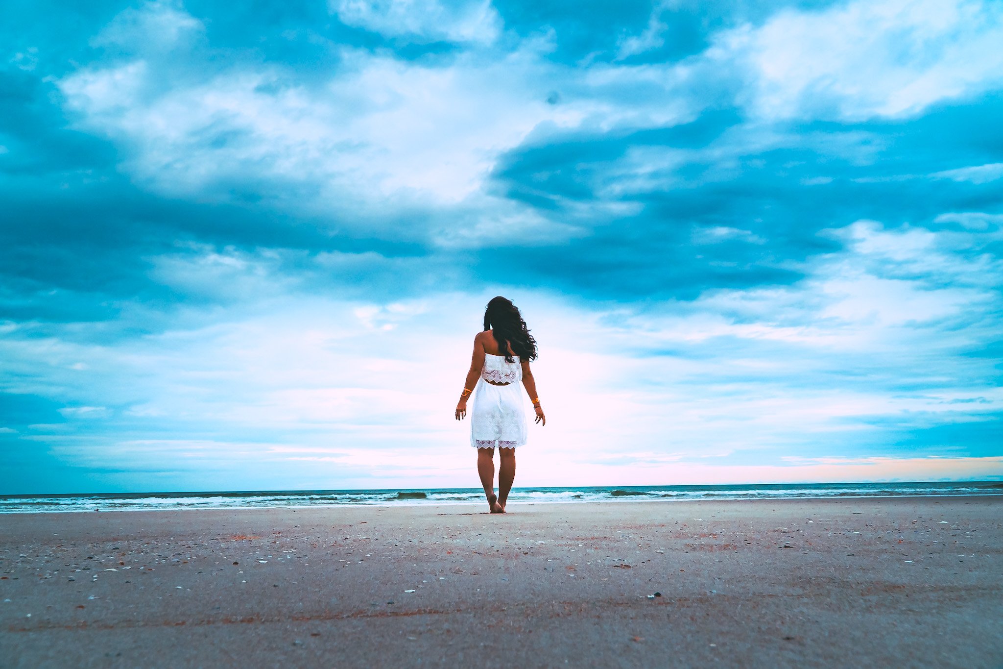 A woman in a white dress stands on a sandy beach facing the ocean with a cloudy sky above.