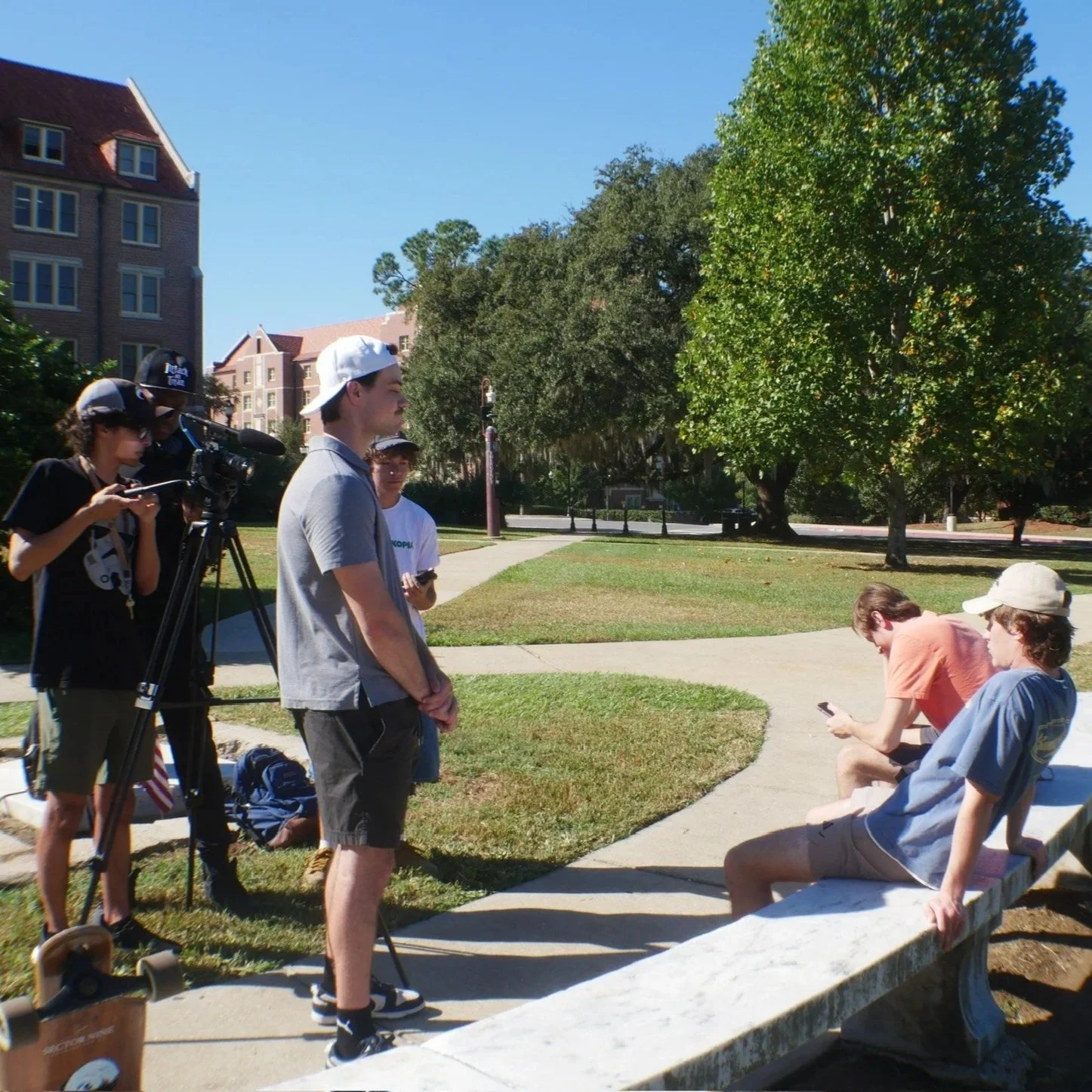 Group of young people filming and taking photos in a park with benches, trees, and buildings in the background on a sunny day.