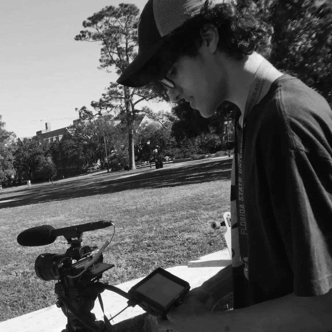 A young man wearing glasses and a hat, standing outdoors in a park, using a smartphone near a mounted camera on a tripod.