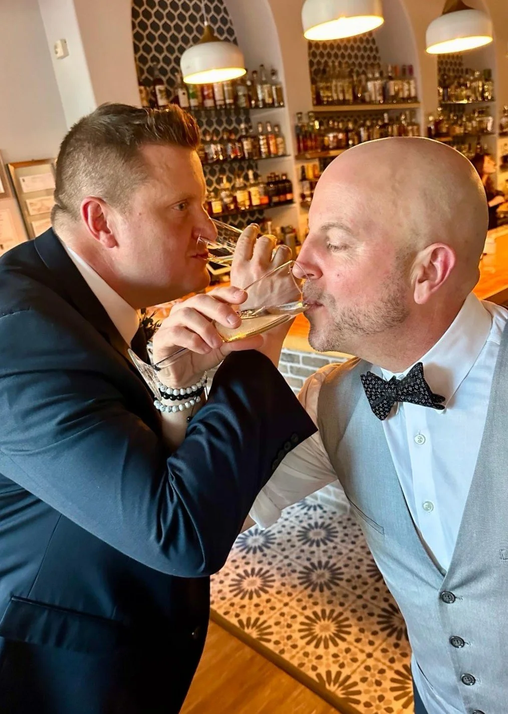 Two men in formal attire, one in a black suit and the other in a light gray vest and white shirt with a bow tie, sharing a drink in a cozy restaurant or bar with shelves of bottles in the background.