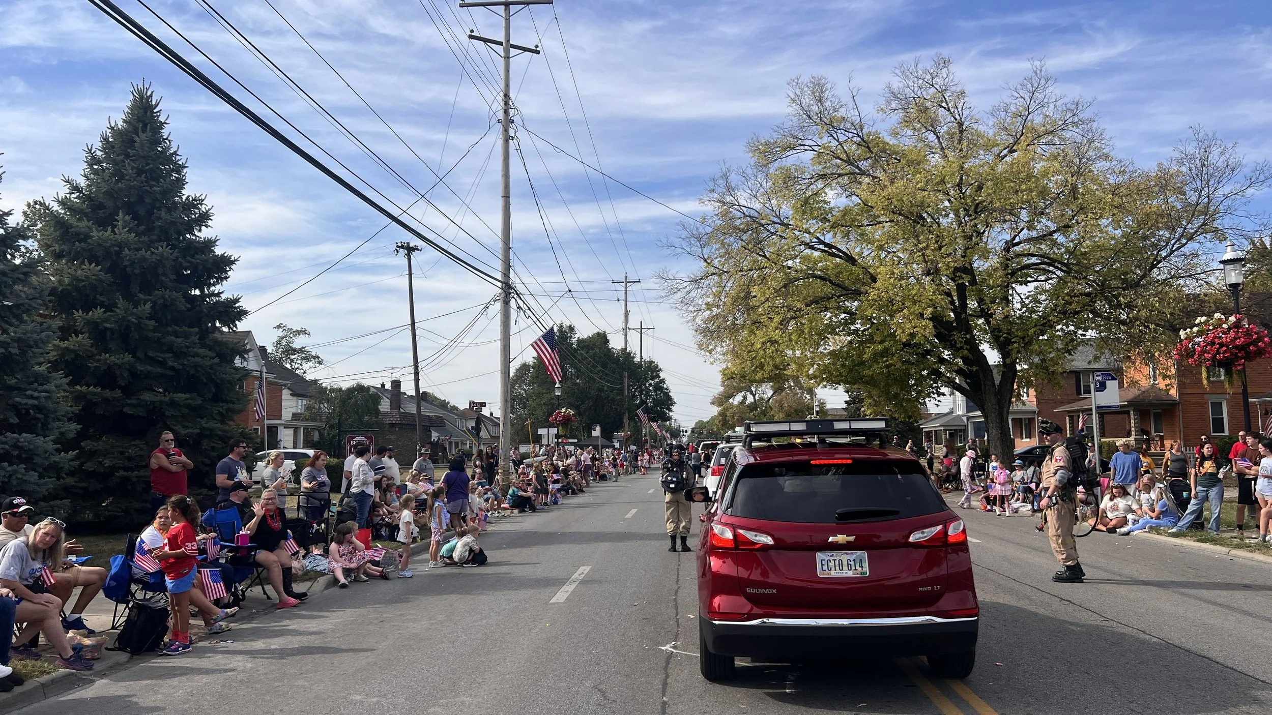 A parade on a small town street with people sitting and standing along the sides, patriotic decorations, and a red Chevrolet SUV driving in the parade. The street is lined with trees, houses, and American flags.