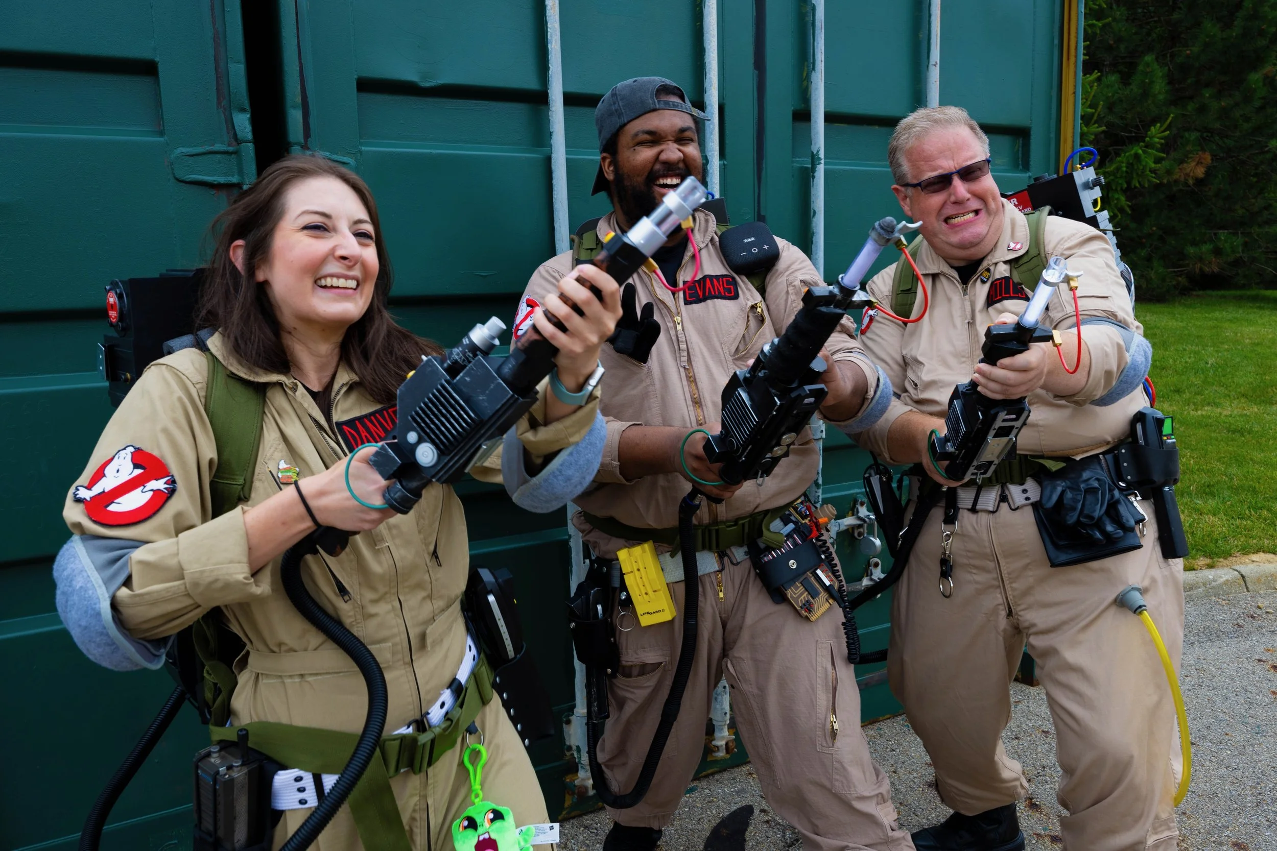 Three people dressed as Ghostbusters holding proton packs and weapons, smiling and laughing outdoors in front of a green shipping container.