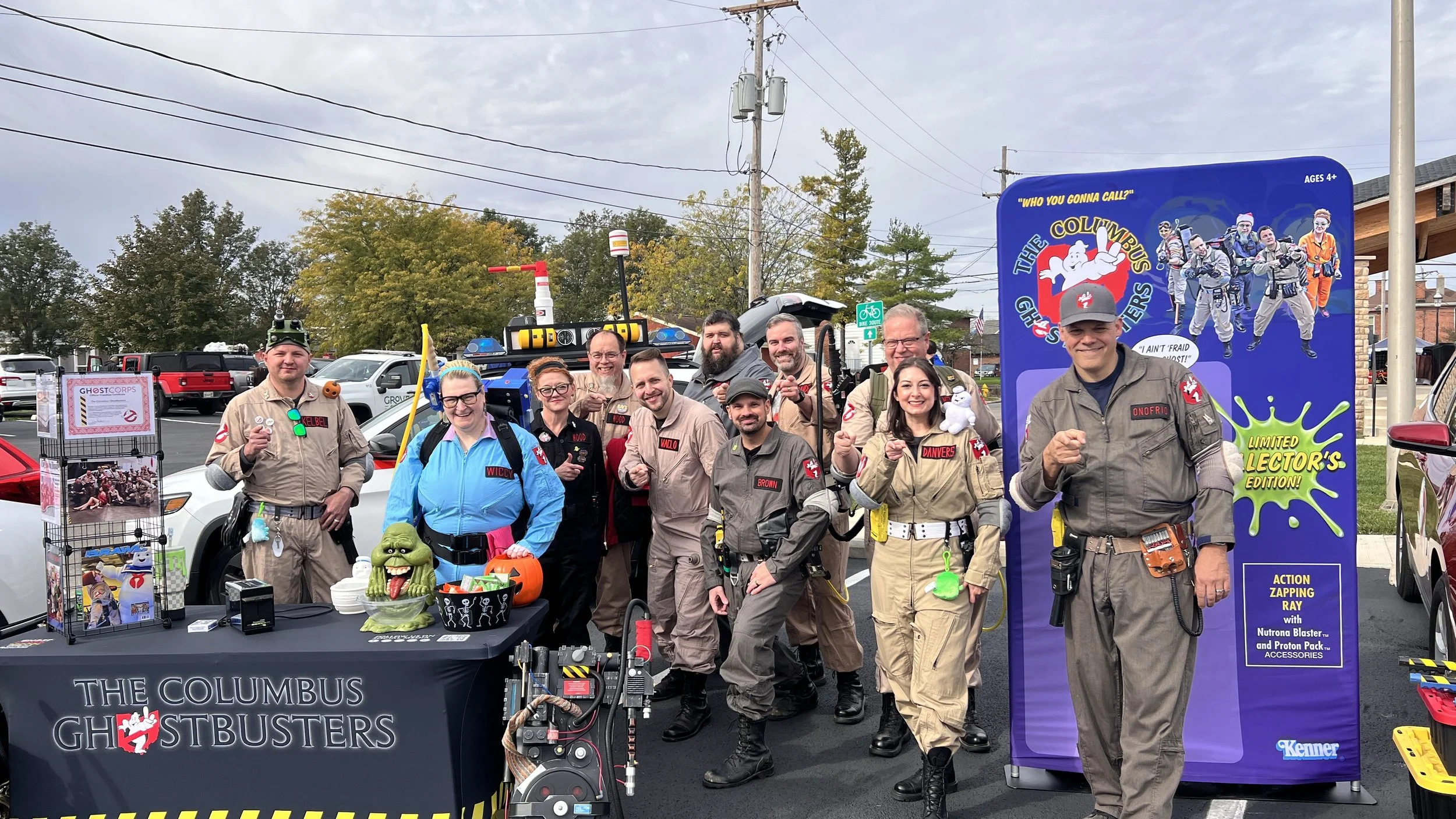 Group of people dressed as Ghostbusters in costumes, standing outdoors in a parking lot, with promotional displays and Ghostbusters-themed accessories.
