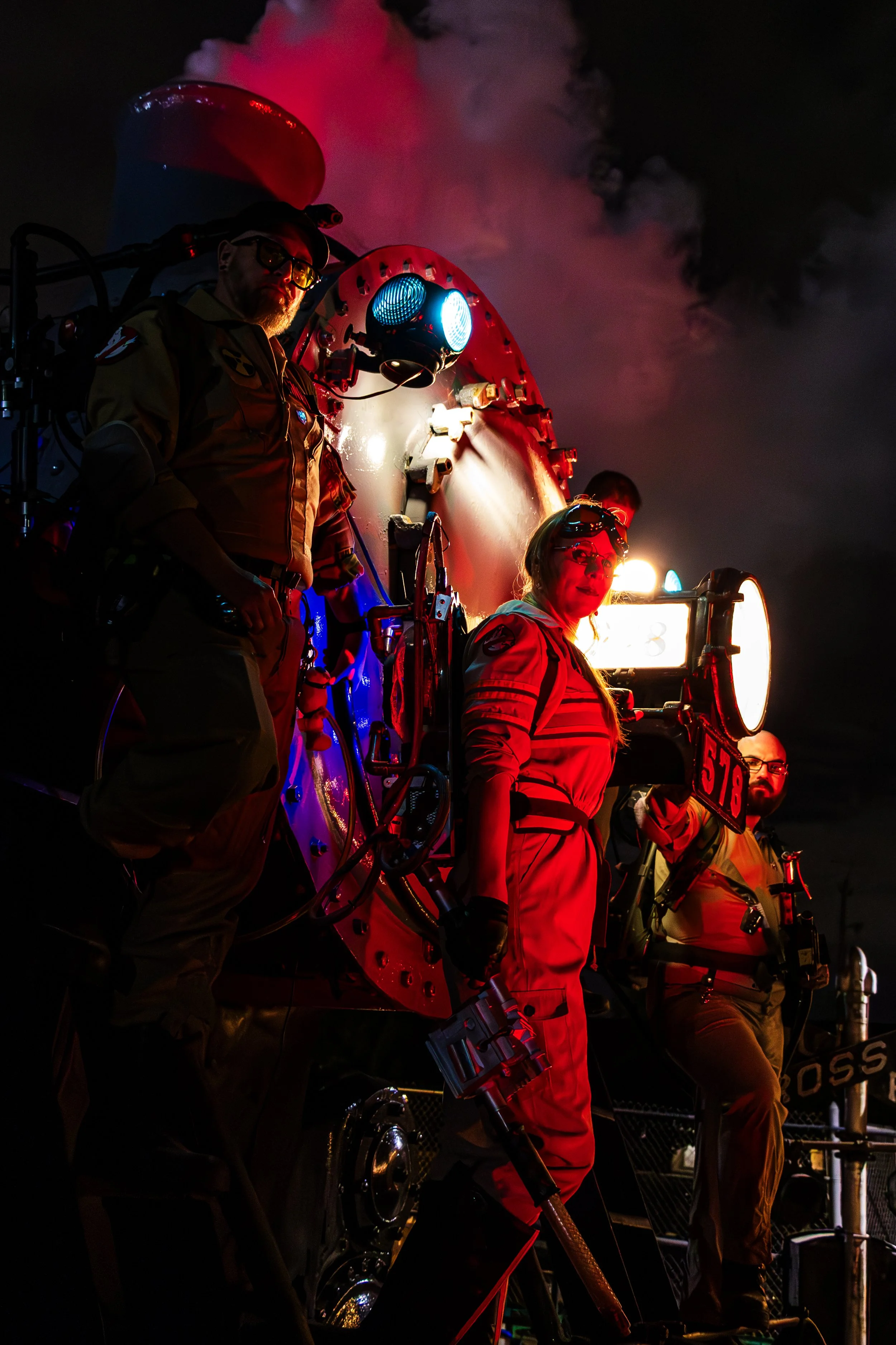 Nighttime scene with three people in astronaut suits, standing on a landing platform, with a large spacecraft emitting purple and red glow and smoke in the background.