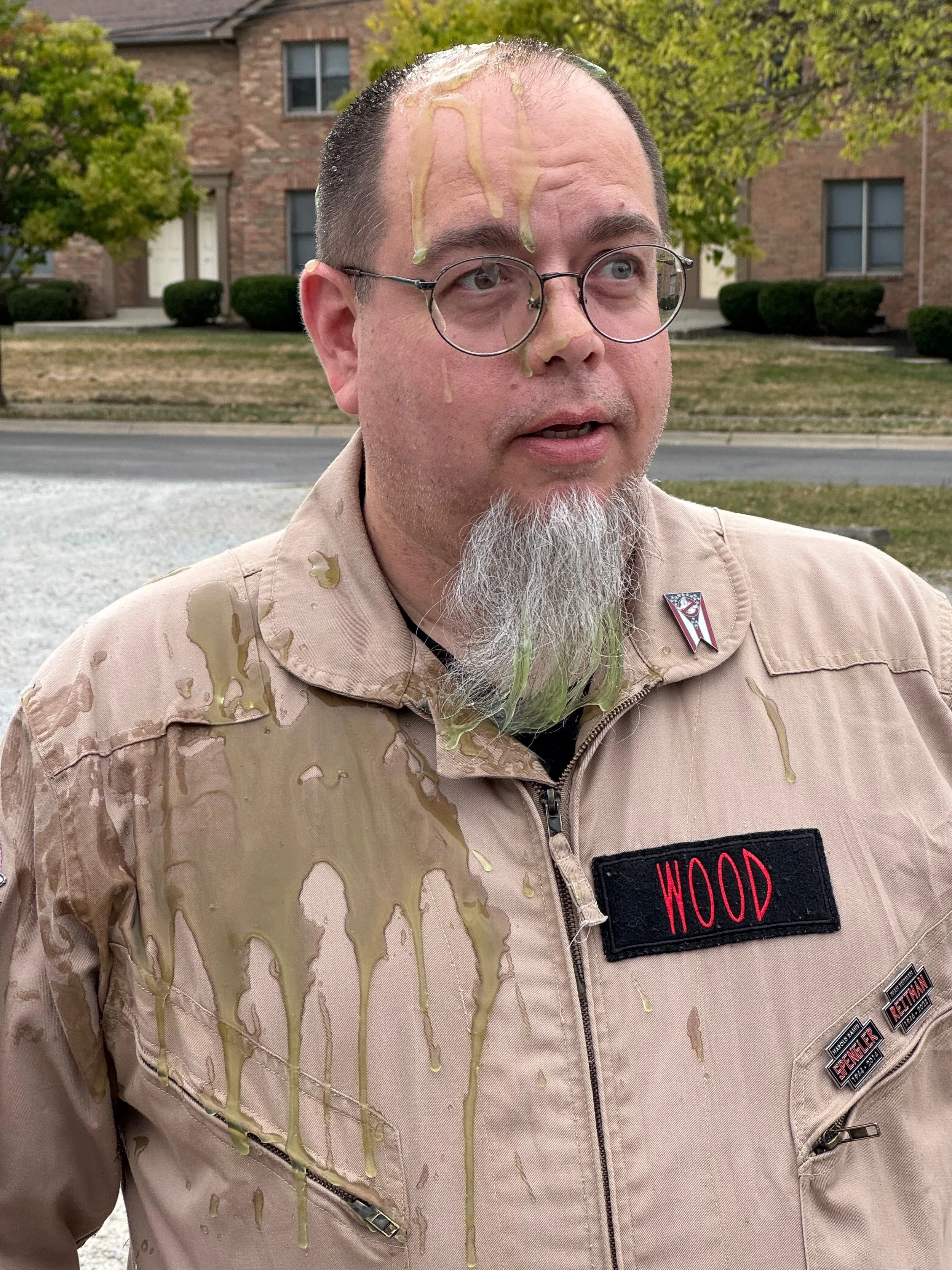 A man with glasses and a beard stands outdoors in front of trees and brick buildings. He is wearing a Ghostbusters uniform with the name tag 'WOOD' and is covered in green slime, on his face and jacket.