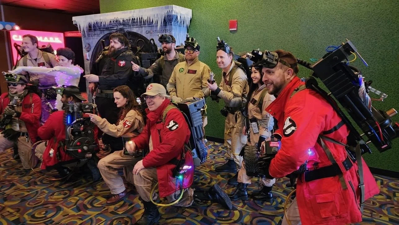 Group of people dressed as Ghostbusters in costumes with proton packs, posing for a photo at a movie premiere.