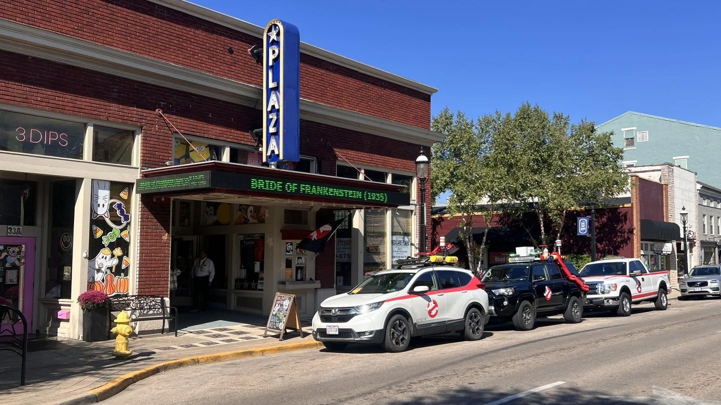 Ghostbusters themed vehicles parked in front of a movie theater with a vertical blue sign that reads 'PLAZA' and a green scrolling marquee. The yellow fire hydrant is near the sidewalk under a clear blue sky.