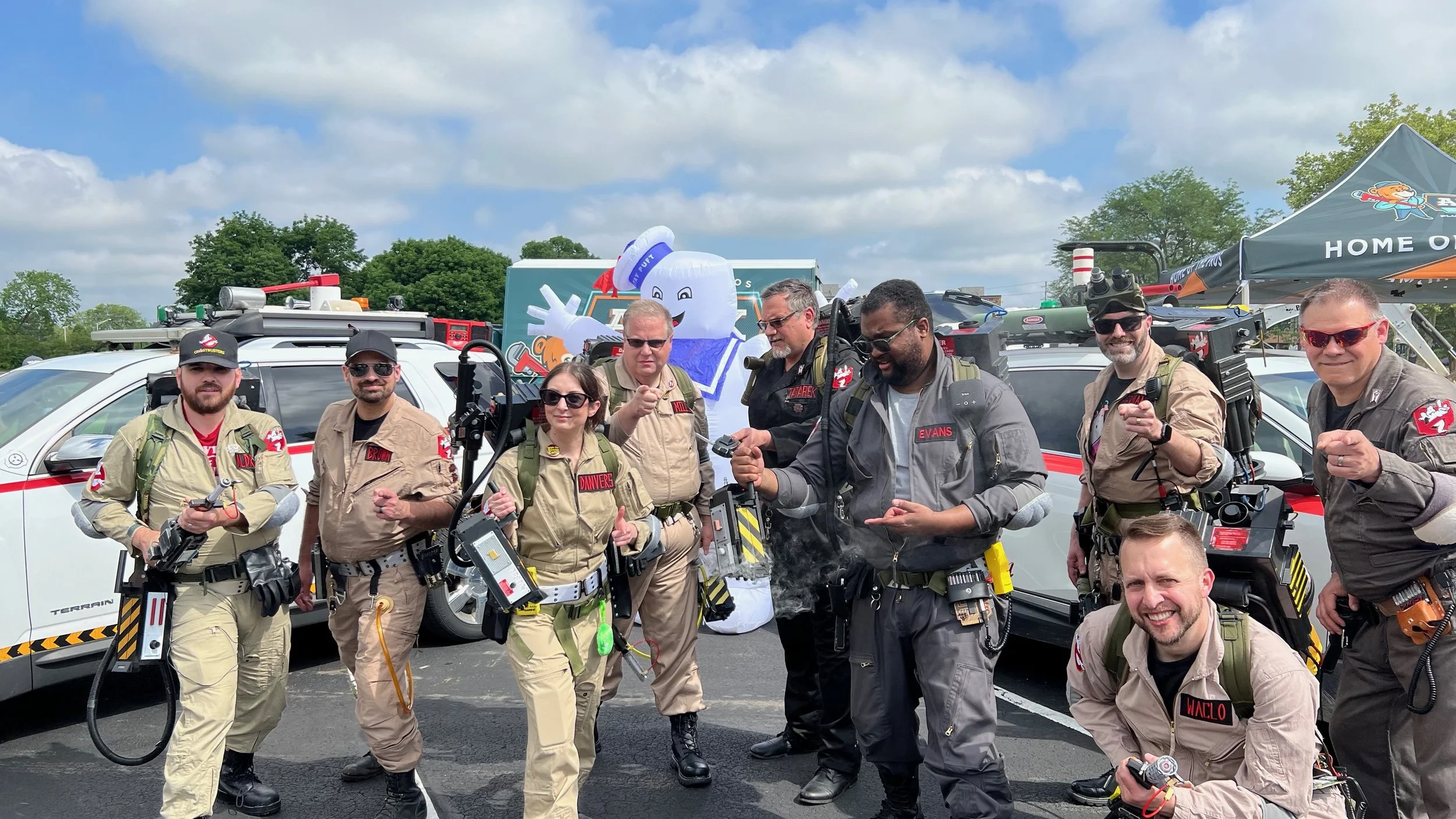 Group of team members dressed in Ghostbusters costumes with proton packs, standing in a parking lot with Ghostbusters-themed tents and a Marshmallow Man mascot in the background.