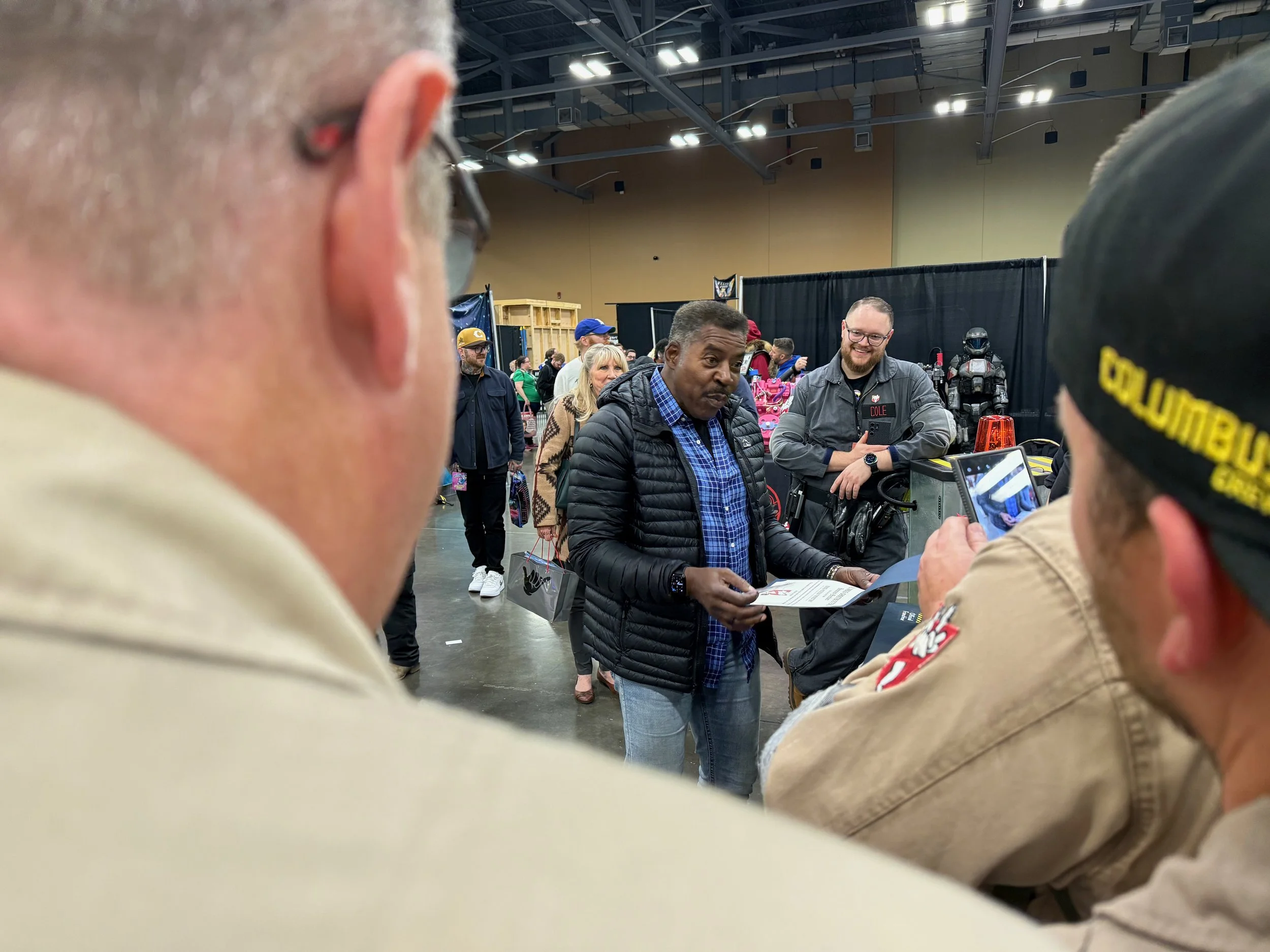 Ernie Hudson holds papers and is speaking to a group of people at GalaxyCon, with colorful booths and attendees in the background. Two men in uniform are visible.