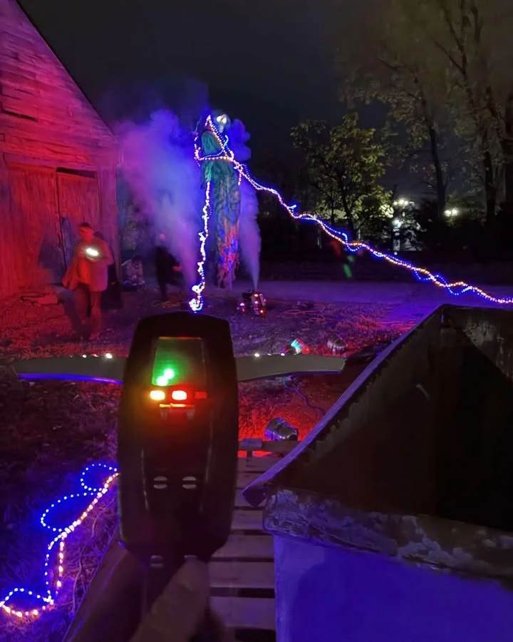 Nighttime scene with people and sheep in a rural setting decorated with colorful Christmas or holiday lights, smoke, and a person holding a handheld device with green and red lights, surrounding a festive display.