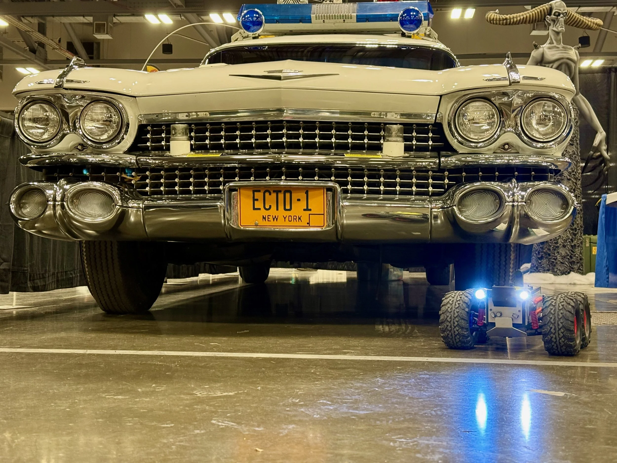Front view of a replica of Ecto-1, displayed at an indoor exhibition with a replica of the RTV with bright headlights in front.