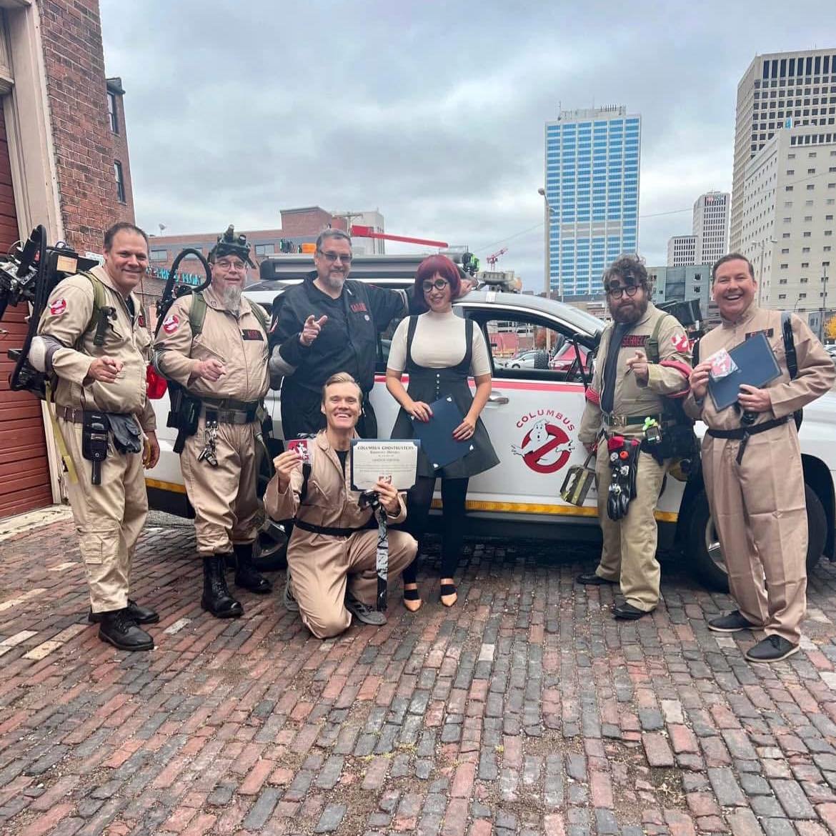 Group of people dressed as Ghostbusters characters standing in front of a Ghostbusters-themed vehicle with city buildings in the background. Some are holding certificates and props, smiling at the camera.