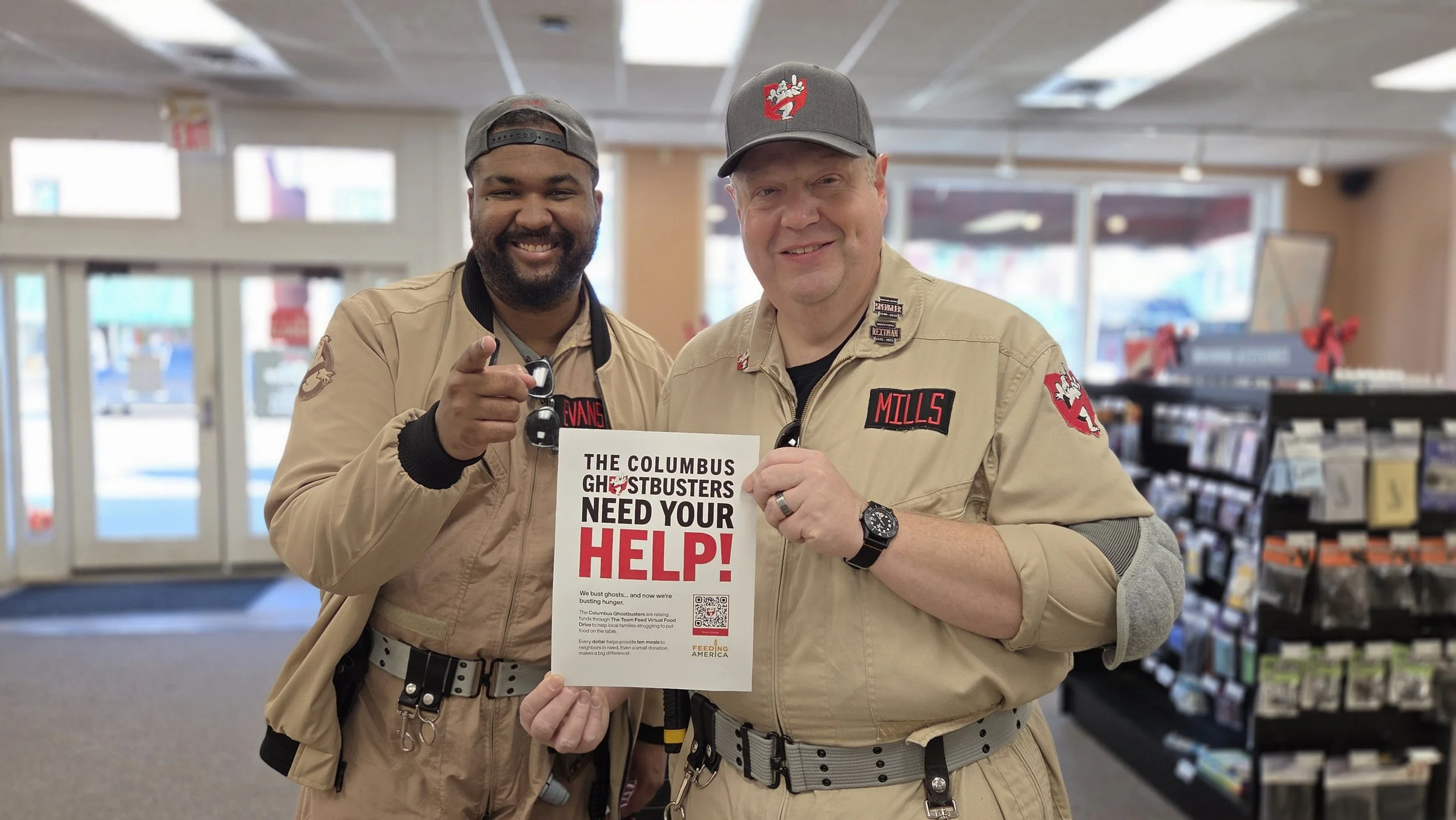 Two men in Ghostbusters costumes standing inside a store, holding a sign that says 'The Columbus Ghostbusters Need Your Help!' with a QR code. They are smiling and pointing at the camera.