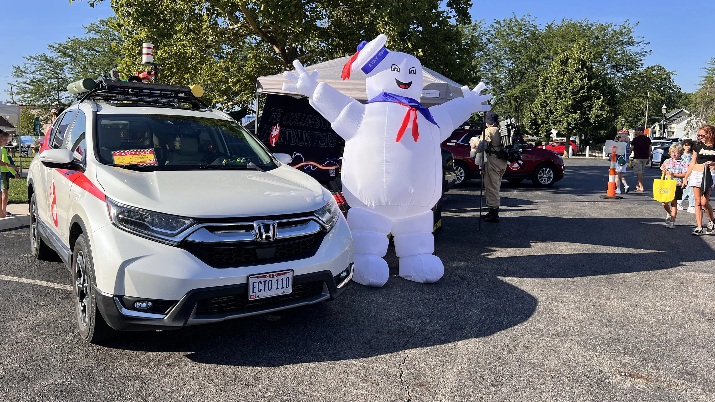 A White Honda car with Ghostbusters decals and a caution sign in the windshield, parked next to an inflatable Stay Puft Marshmallow Man at a busy outdoor event with people walking and trees in the background.