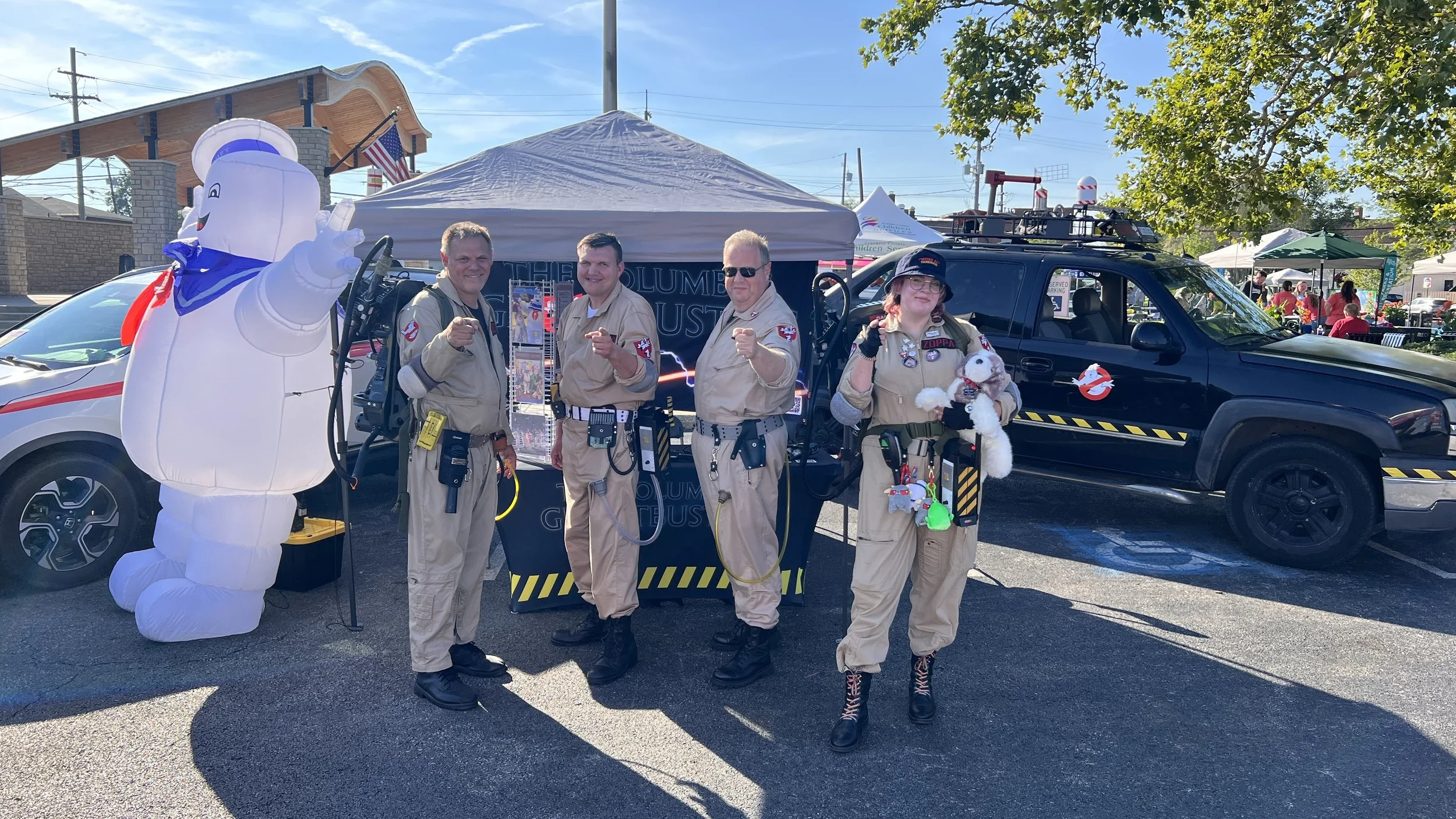 Group of four people in Ghostbusters costumes standing in front of a Ghostbusters-themed display and police vehicle, with a large inflatable Stay Puft Marshmallow Man and a crowd in the background at an outdoor event.