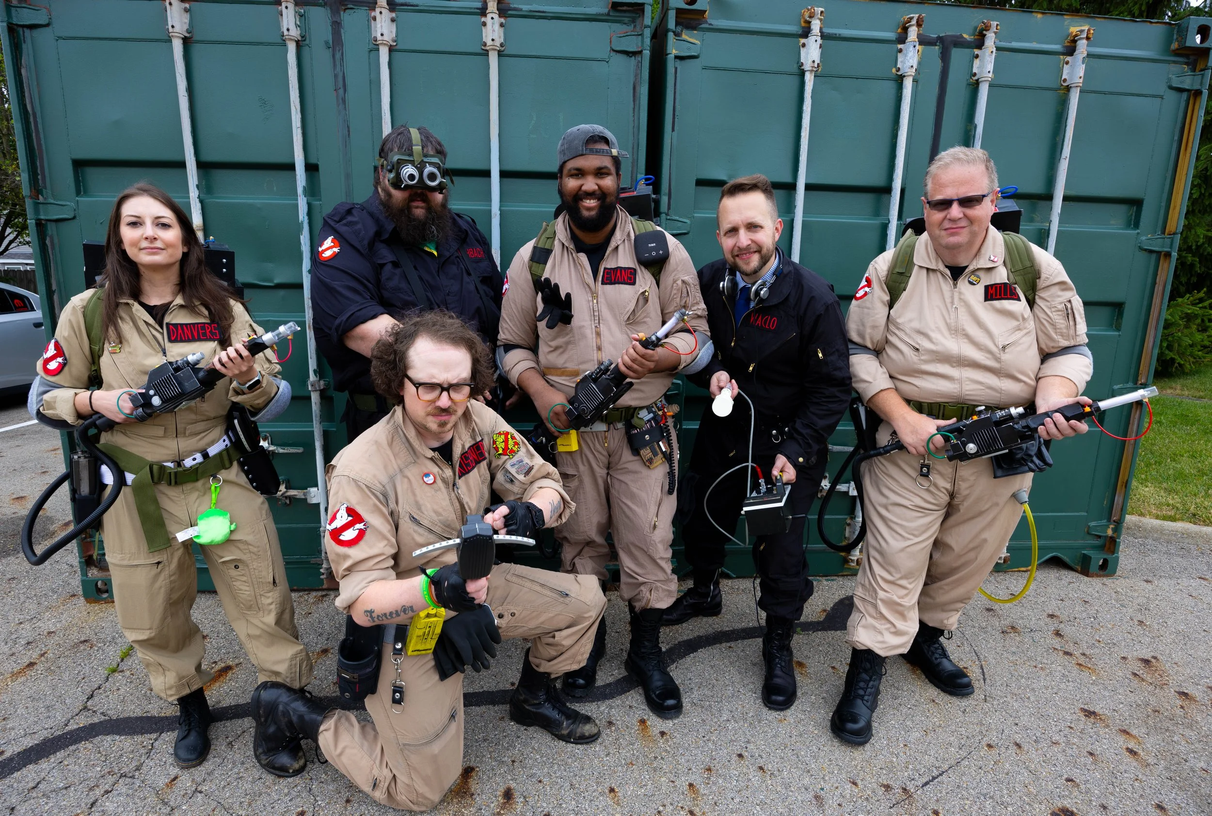 Group of seven people dressed as Ghostbusters in ghostbusting costumes, holding proton packs and ghost traps, standing in front of a green shipping container.