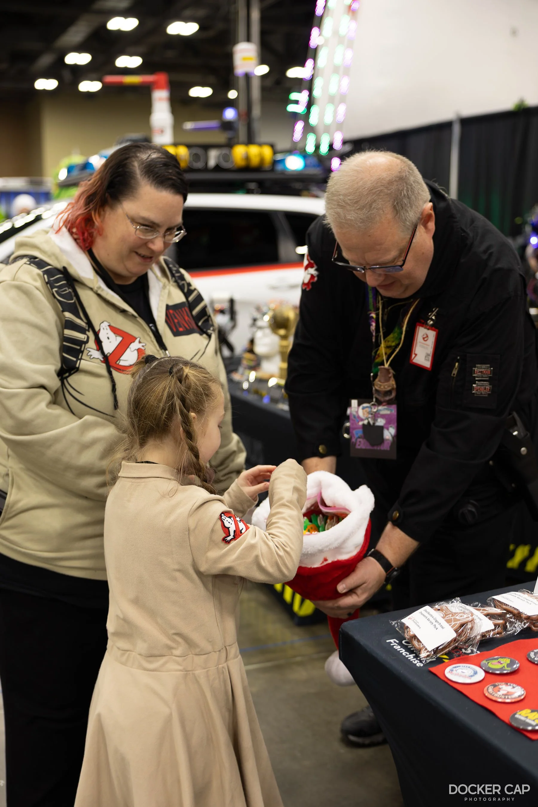 A young girl and two adults dressed in Ghostbusters-themed clothing at a convention, exchanging gifts next to a Ghostbusters vehicle with lights and equipment in the background.