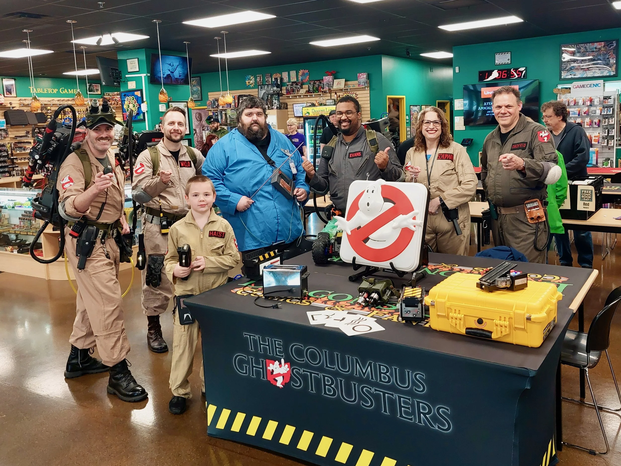 Group of people dressed as Ghostbusters standing around a table with Ghostbusters-themed items inside a store, with a large Ghostbusters logo and display on the table.