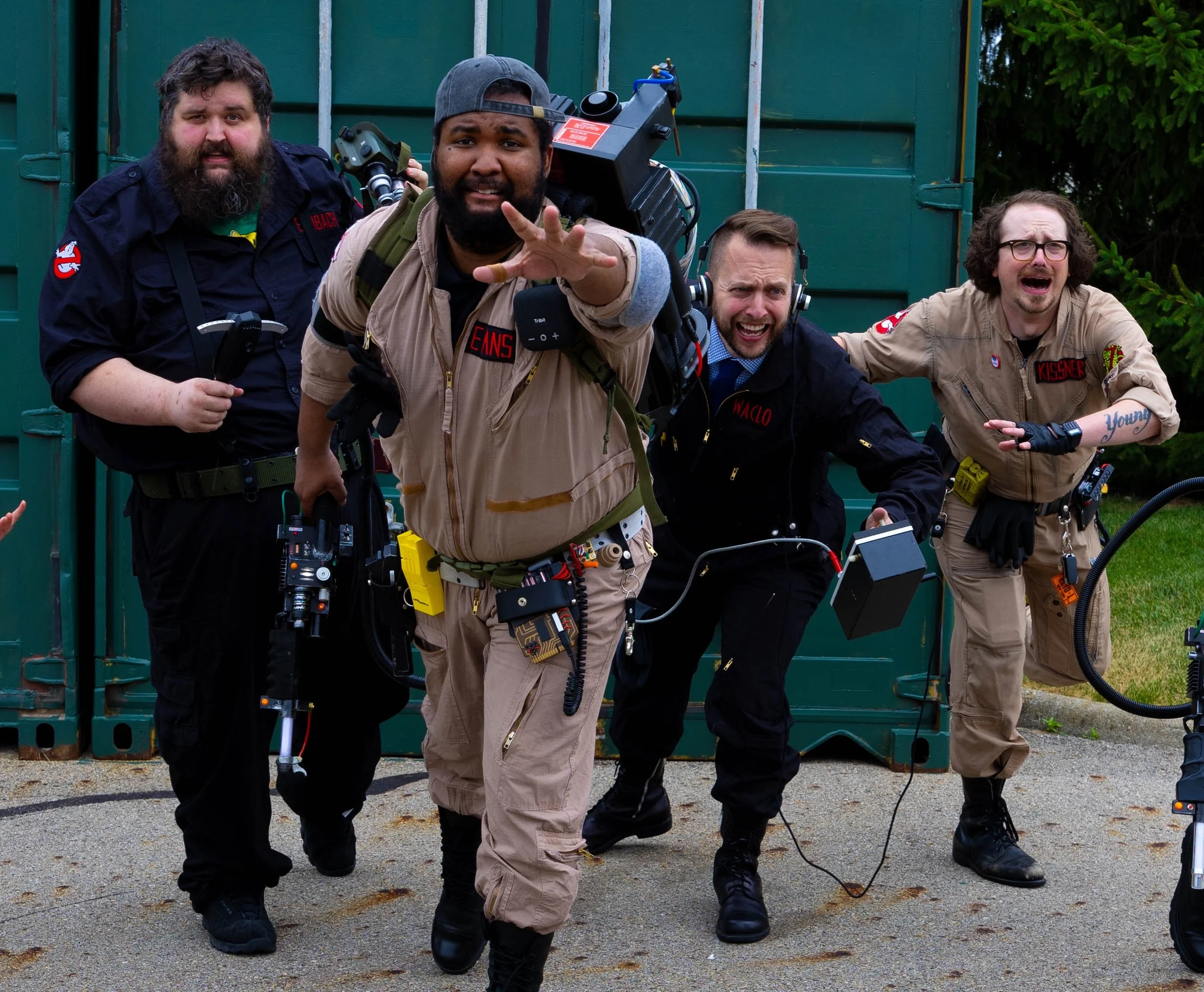 Four men dressed as Ghostbusters running in front of storage containers.