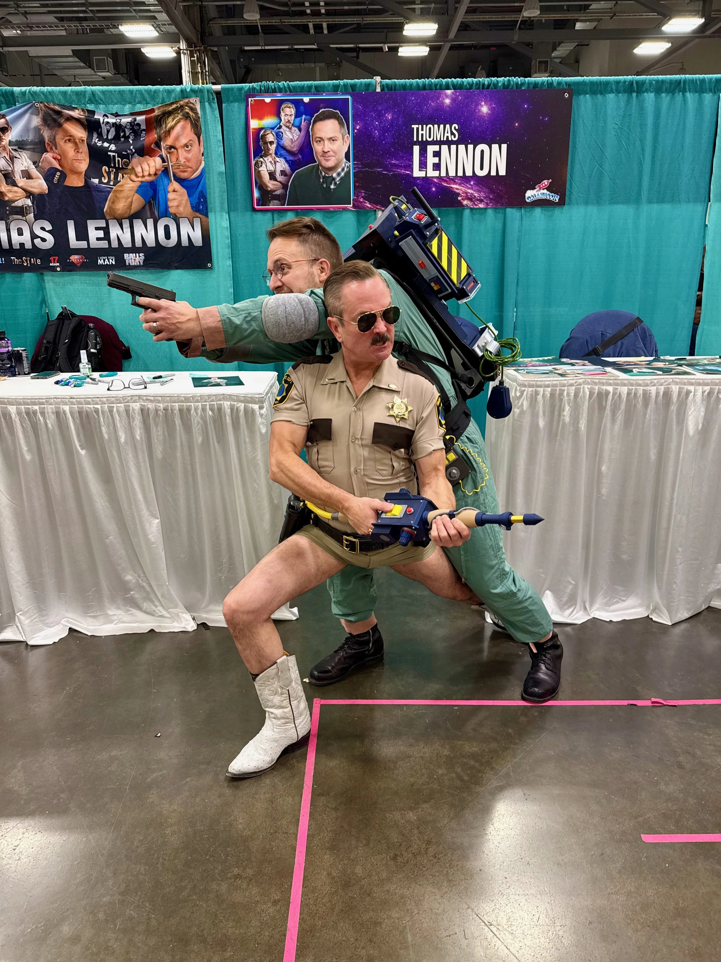 A Ghostbuster and Thomas Lennon dressed as Lieutenant Dangle from Reno 911, posing dynamically at a convention booth with posters of Thomas Lennon in the background.