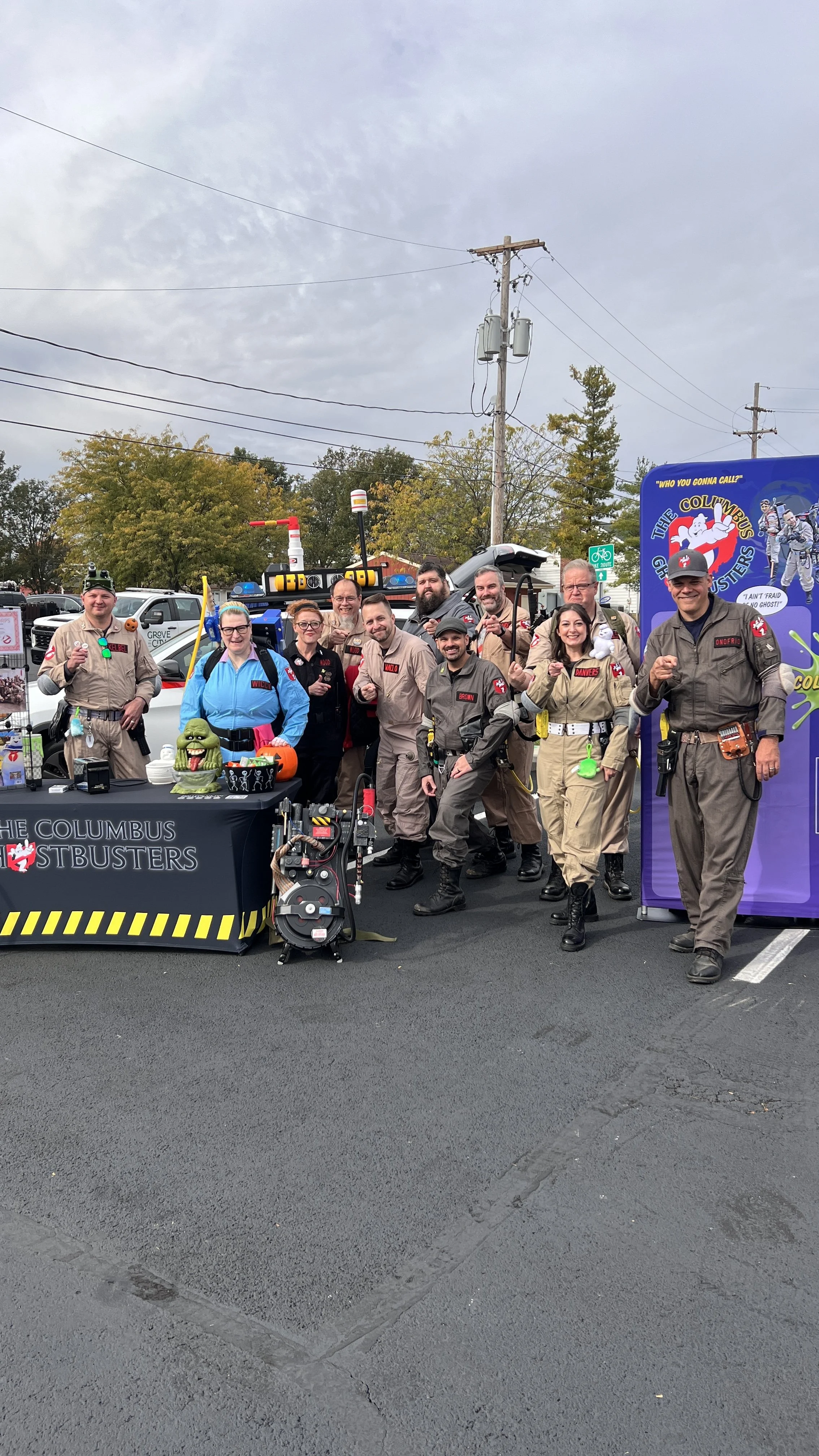 A group of people dressed as Ghostbusters, standing outdoors in a parking lot during daytime, with a display table and photo booth themed for the Ghostbusters movie.