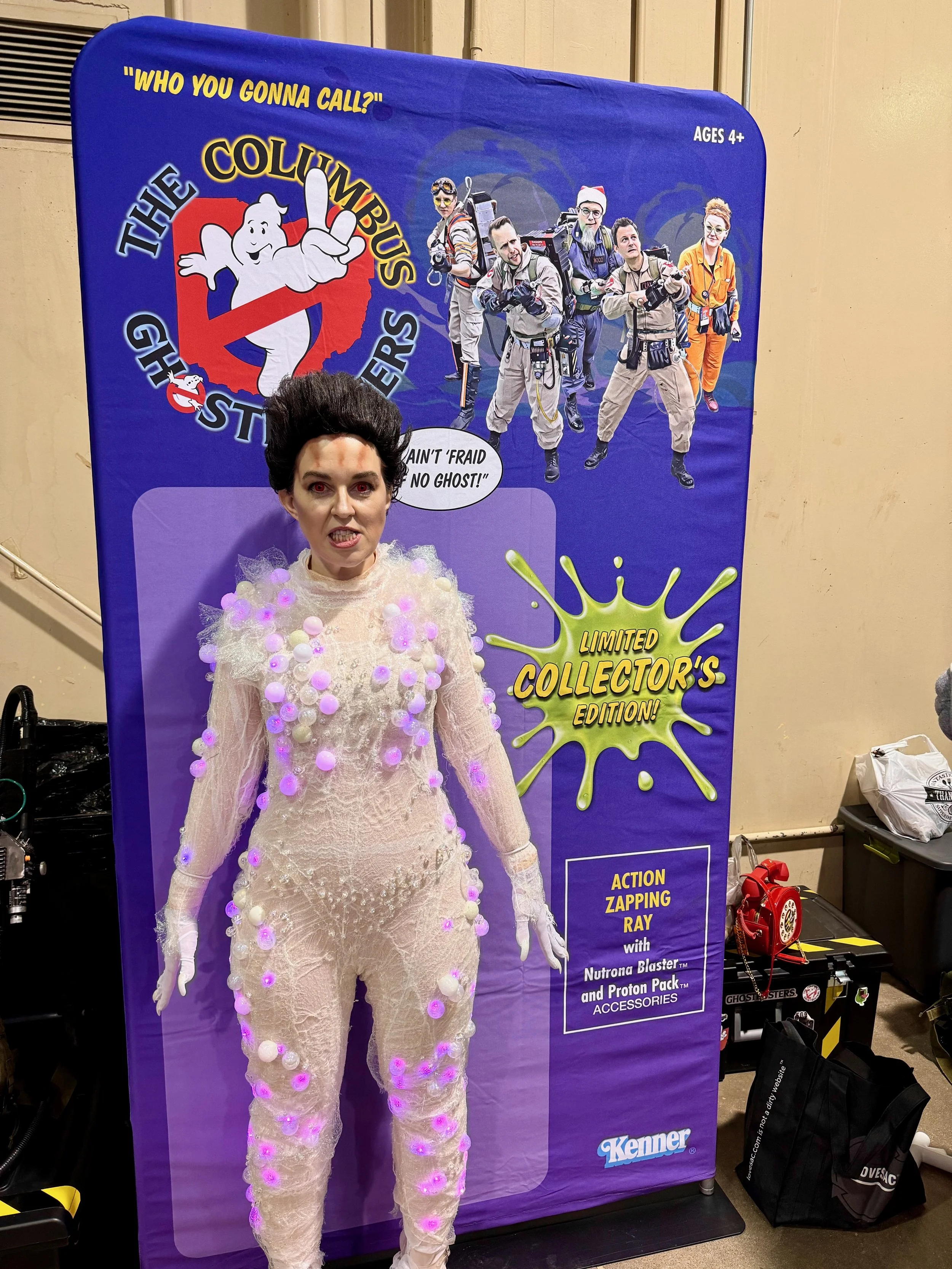 Person dressed as Gozer with glowing pink and purple lights in a promotional display for a Ghostbusters toy line, featuring a large poster of the Ghostbusters team and the slogan 'Who you gonna call?'