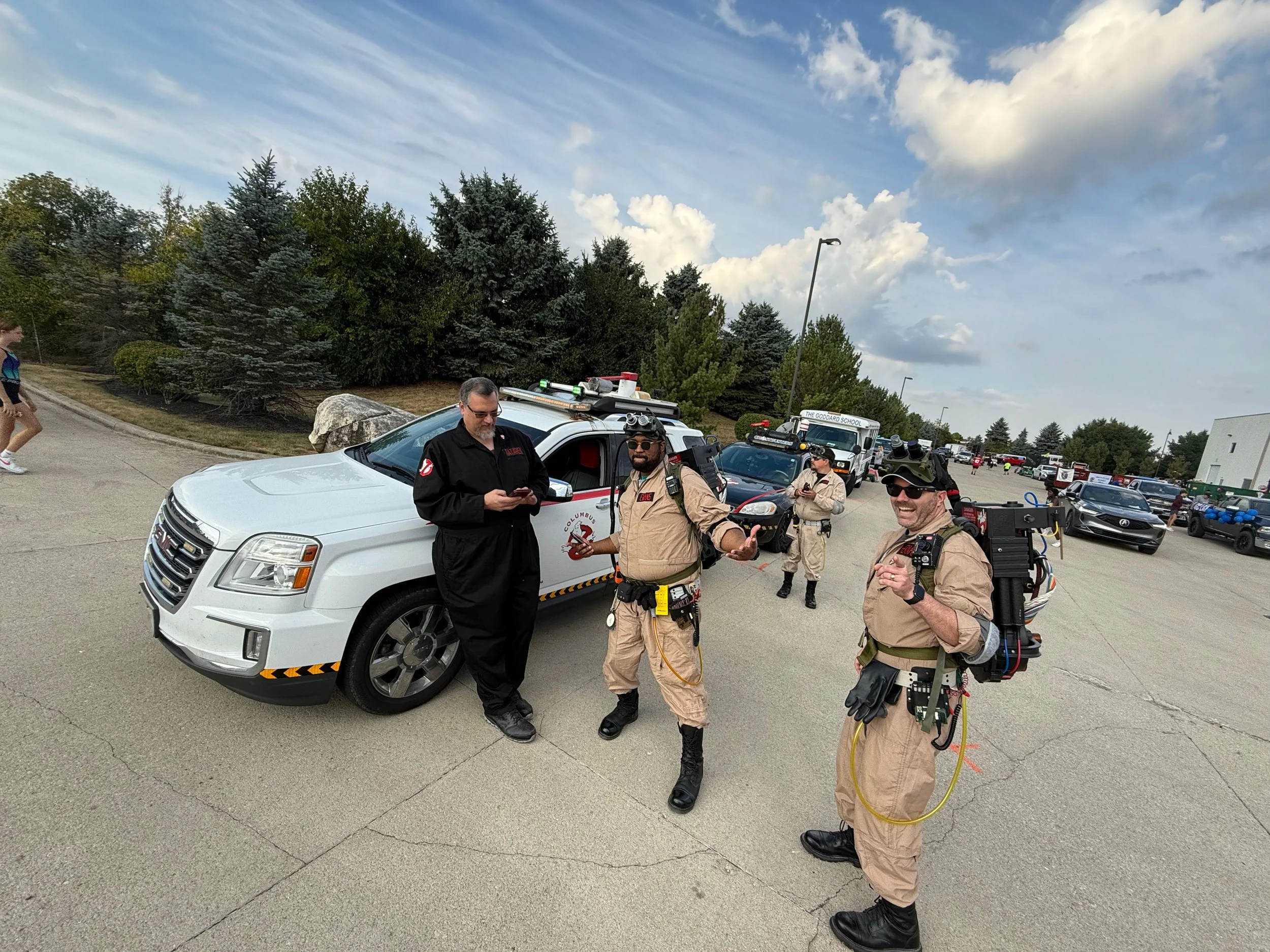 A group of people, some dressed as Ghostbusters, standing near vehicles in a parking lot, with some trees and a partly cloudy sky overhead.