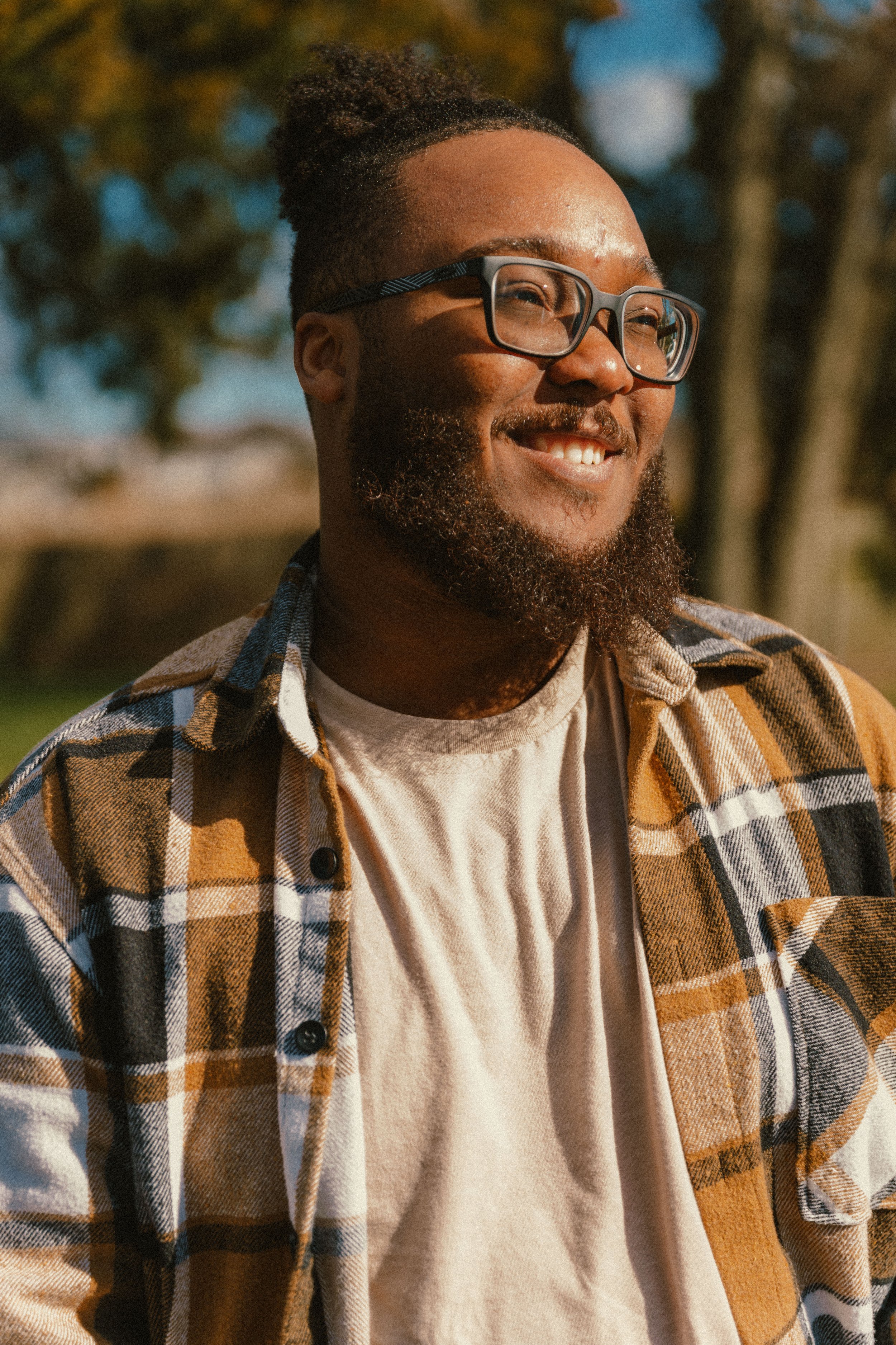 A smiling man with glasses, a beard, and dreadlocks tied back, wearing a plaid shirt over a plain white t-shirt, standing outdoors on a sunny day with trees in the background.