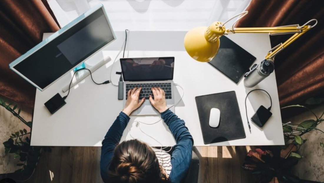 Top-down view of a person working at a cluttered white desk with multiple monitors, a laptop, wireless mouse and mouse pad, water bottle, tablet, and a yellow adjustable desk lamp.