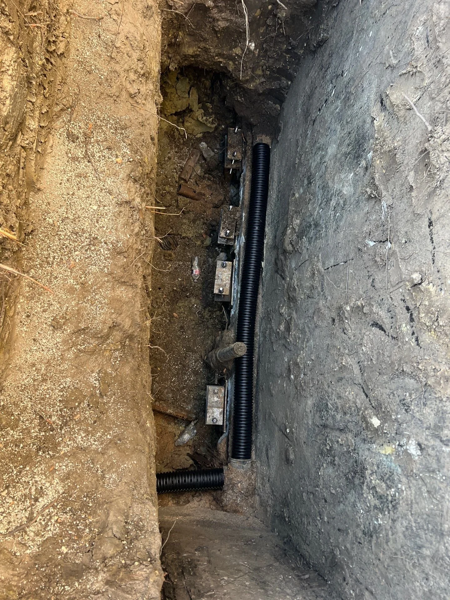 Underground utility conduit installation with black corrugated tubing and metal brackets in a narrow trench.