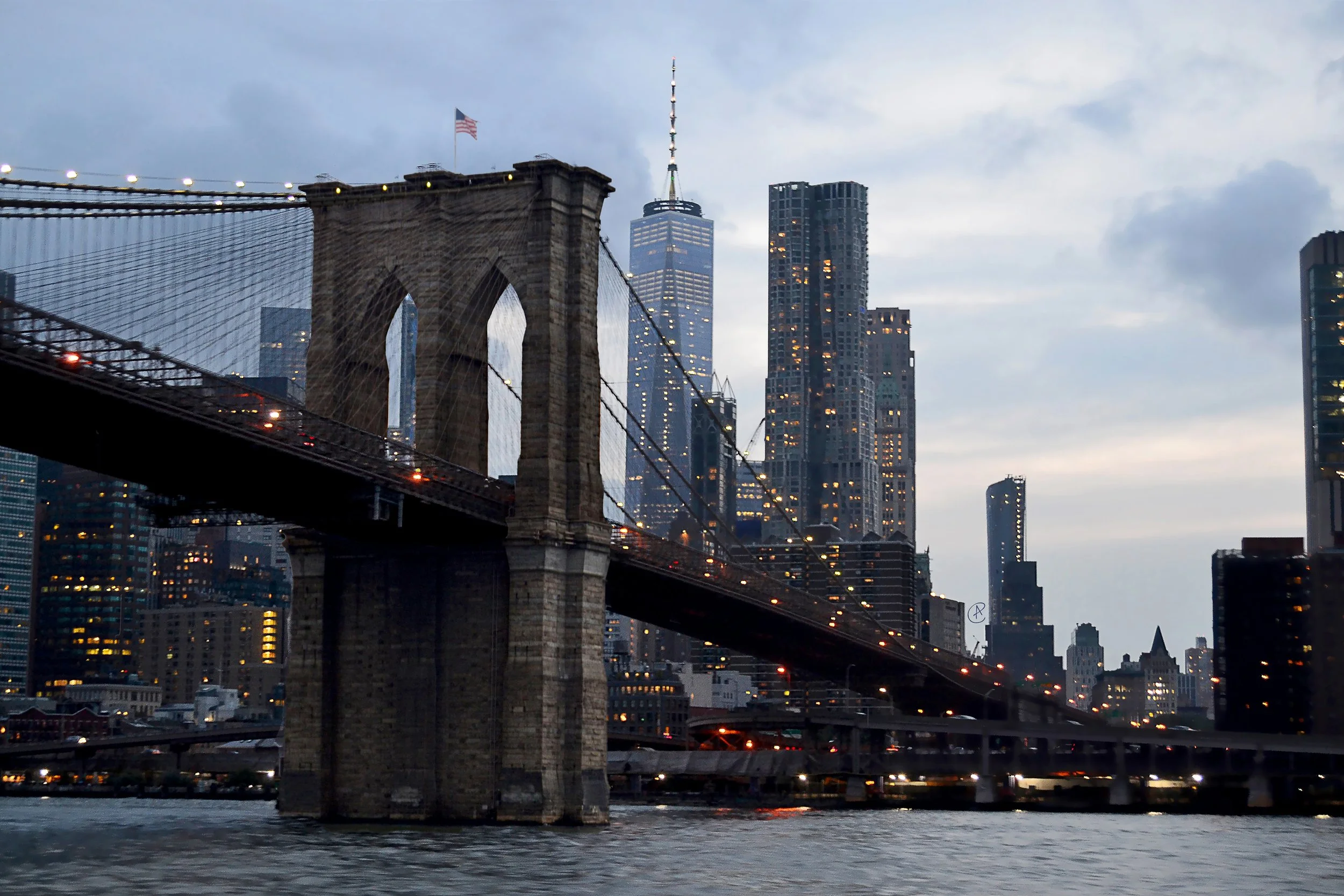 New York City skyline featuring the Brooklyn Bridge with tall skyscrapers, including One World Trade Center, under a cloudy sky at dusk