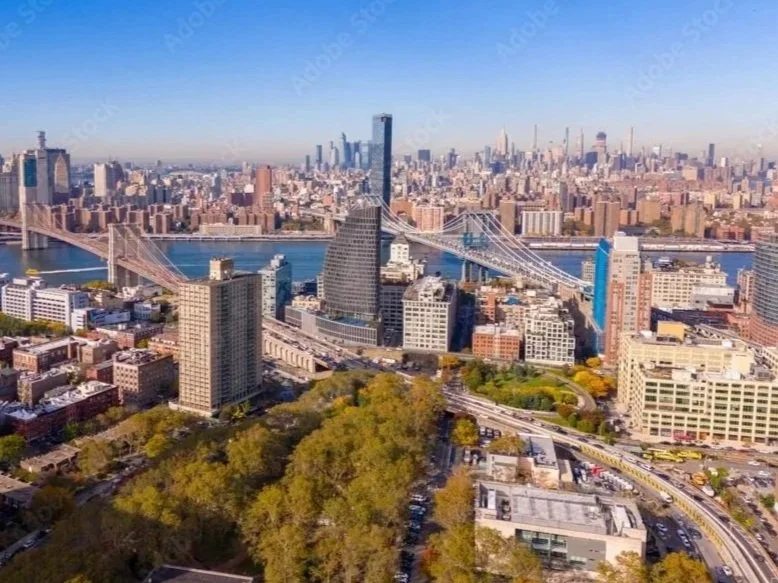 Aerial view of New York City skyline featuring the Brooklyn Bridge, skyscrapers, and the Manhattan Bridge over the East River in the background under a clear blue sky.