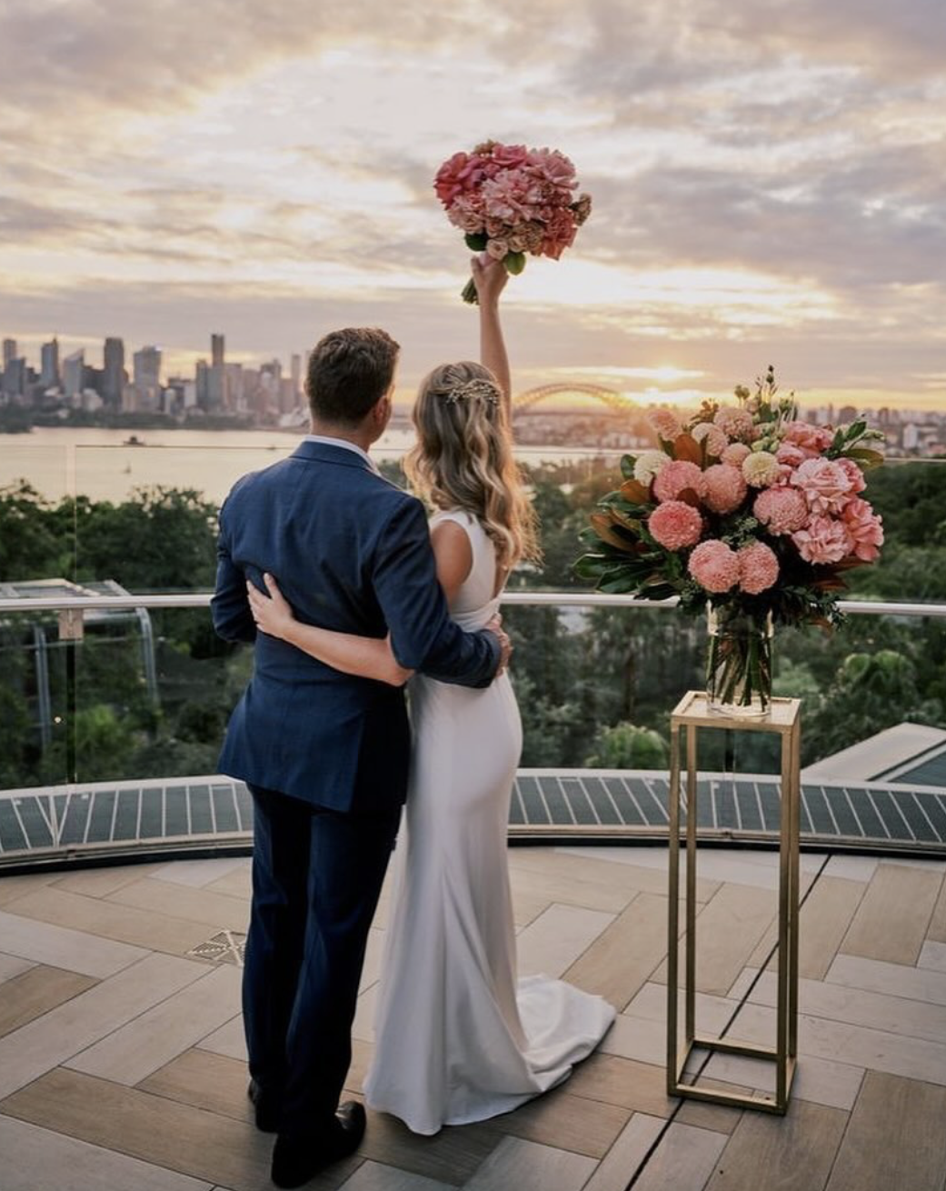 A bride and groom embrace on a terrace during sunset, with ocean and city skyline in the background. The bride is holding a large pink flower bouquet above her head, and a vase of pink flowers is on a tall stand nearby.