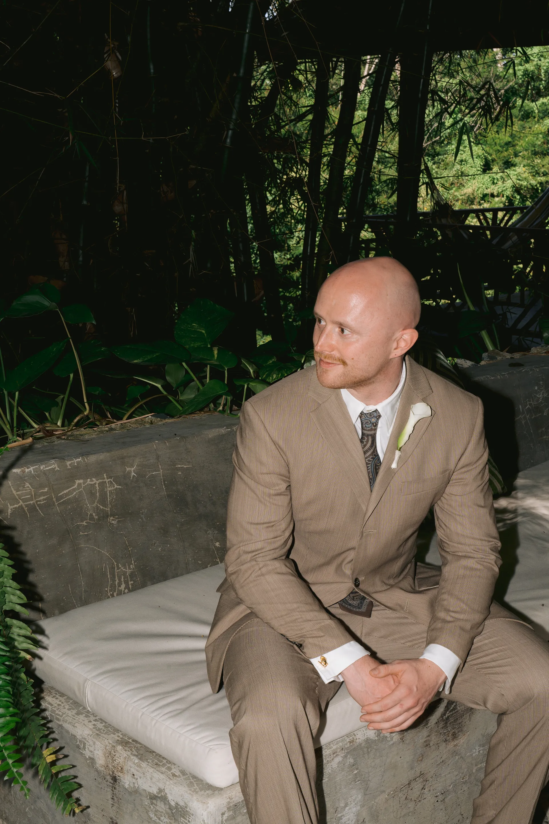 A man in a tan suit with a white shirt and a floral tie sitting on a cushioned bench outdoors, surrounded by tropical plants and dense foliage, with a flower boutonniere pinned to his lapel.