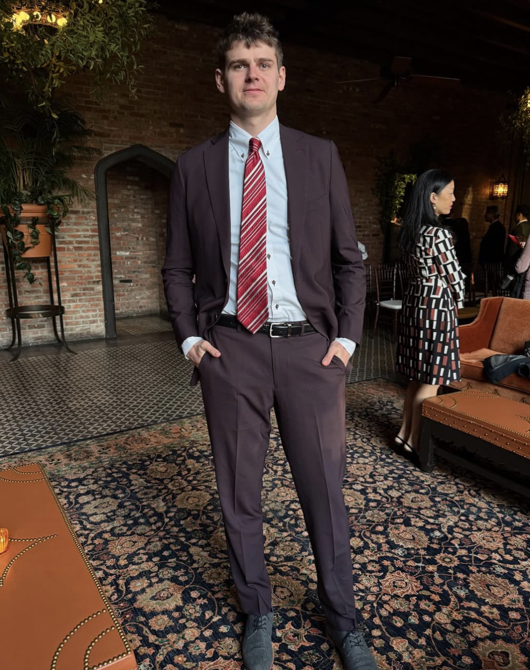 A young man in a purple suit standing with hands in pockets in a dimly lit room with brick walls and patterned carpet, with people in the background at a social gathering.