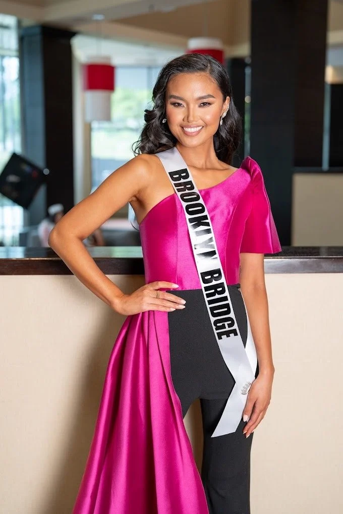 A young woman standing indoors, wearing a bright pink dress with a sash that reads "Brooklyn Bridge". She is smiling, with one hand on her hip, and has shoulder-length dark wavy hair.