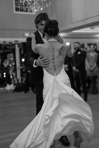 A couple dancing closely at a formal event, with the woman wearing a white gown and the man in a dark suit; there are onlookers in the background.