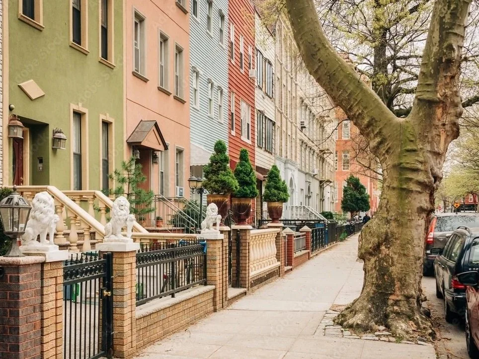 A city sidewalk lined with colorful row houses, large potted trees, a sturdy fence topped with lion statues, and a large tree with expansive branches.