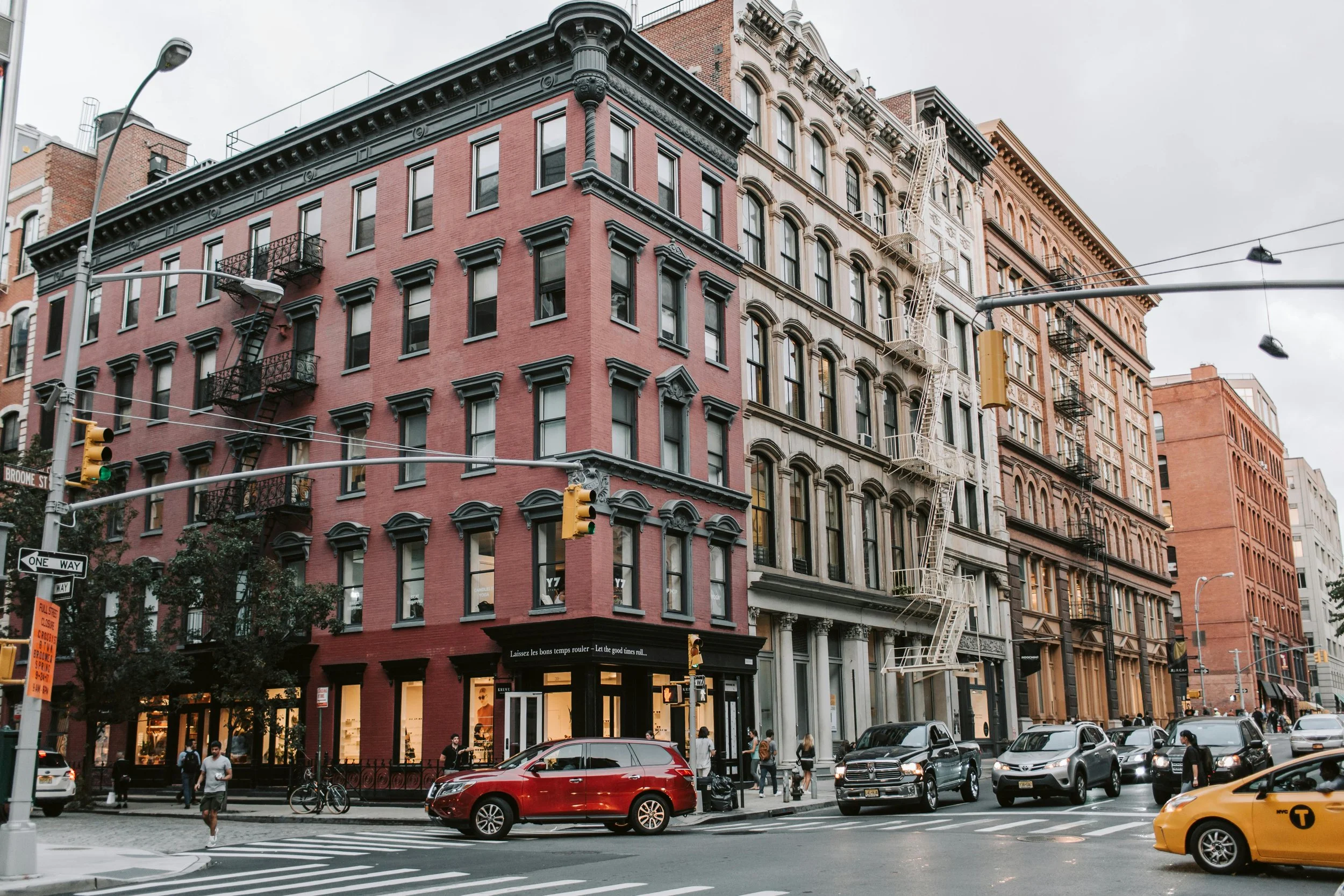 City street scene with a pink and beige brick-building, storefront on the ground level, cars waiting at a red light, pedestrians crossing, and fire escapes on the building.