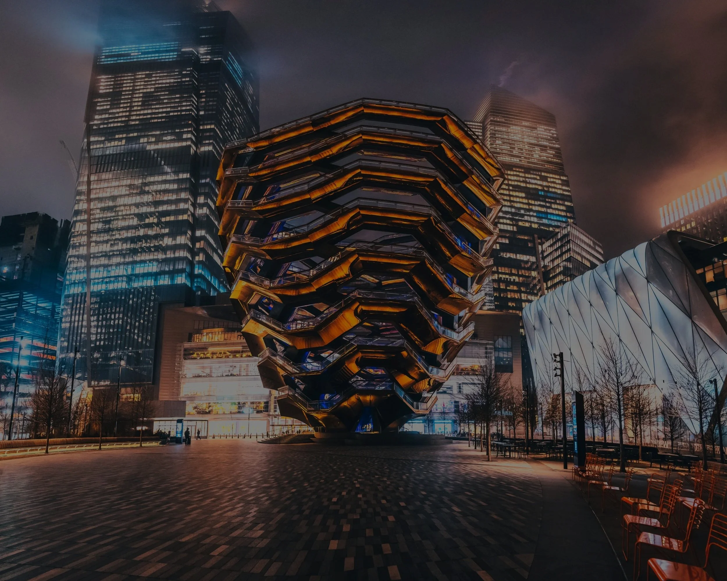 Nighttime cityscape with modern skyscrapers, including the Vessel structure in New York City surrounded by benches and trees.