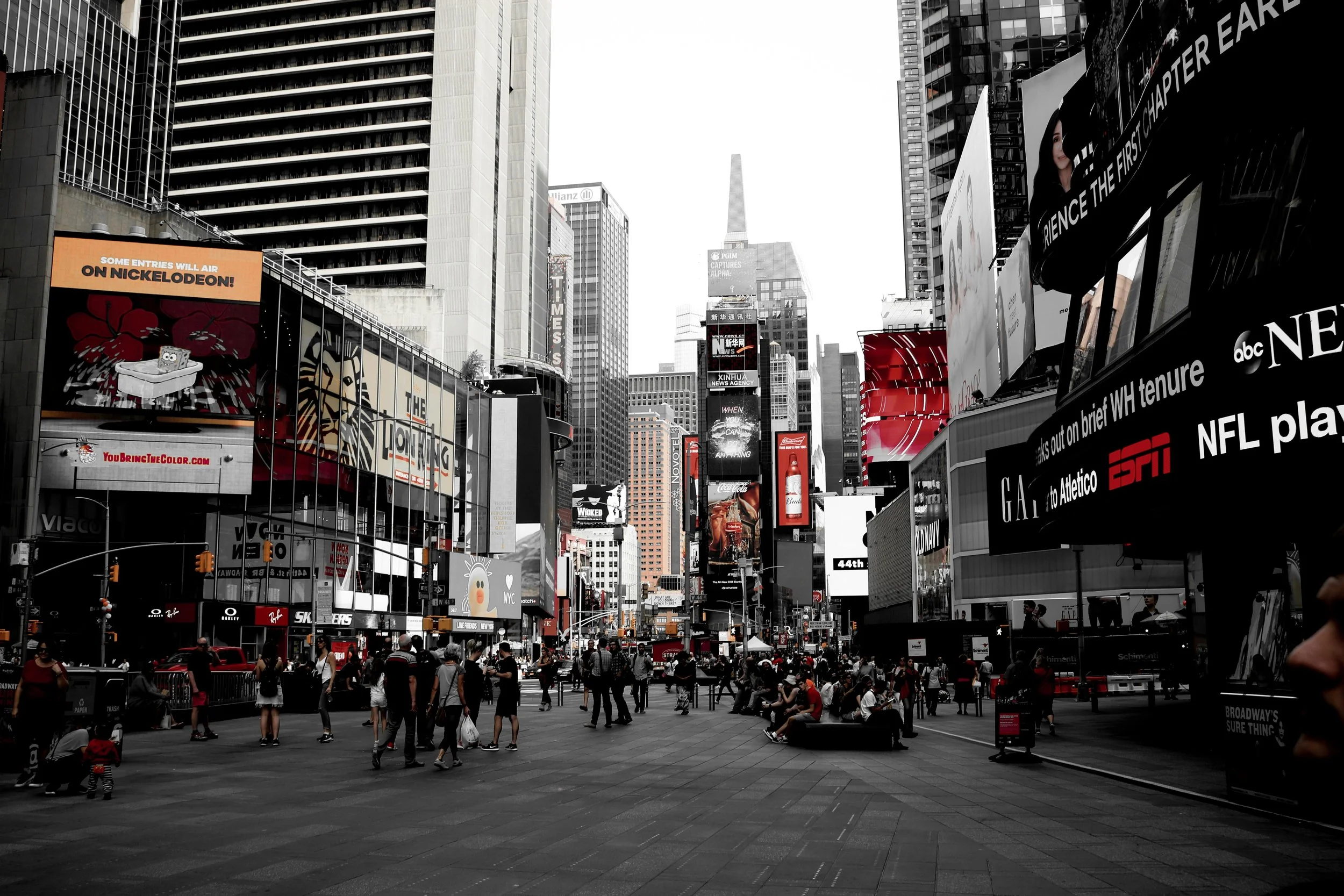 A busy city square in Times Square, New York City, with numerous people walking and sitting, surrounded by tall buildings and large digital billboards displaying ads.
