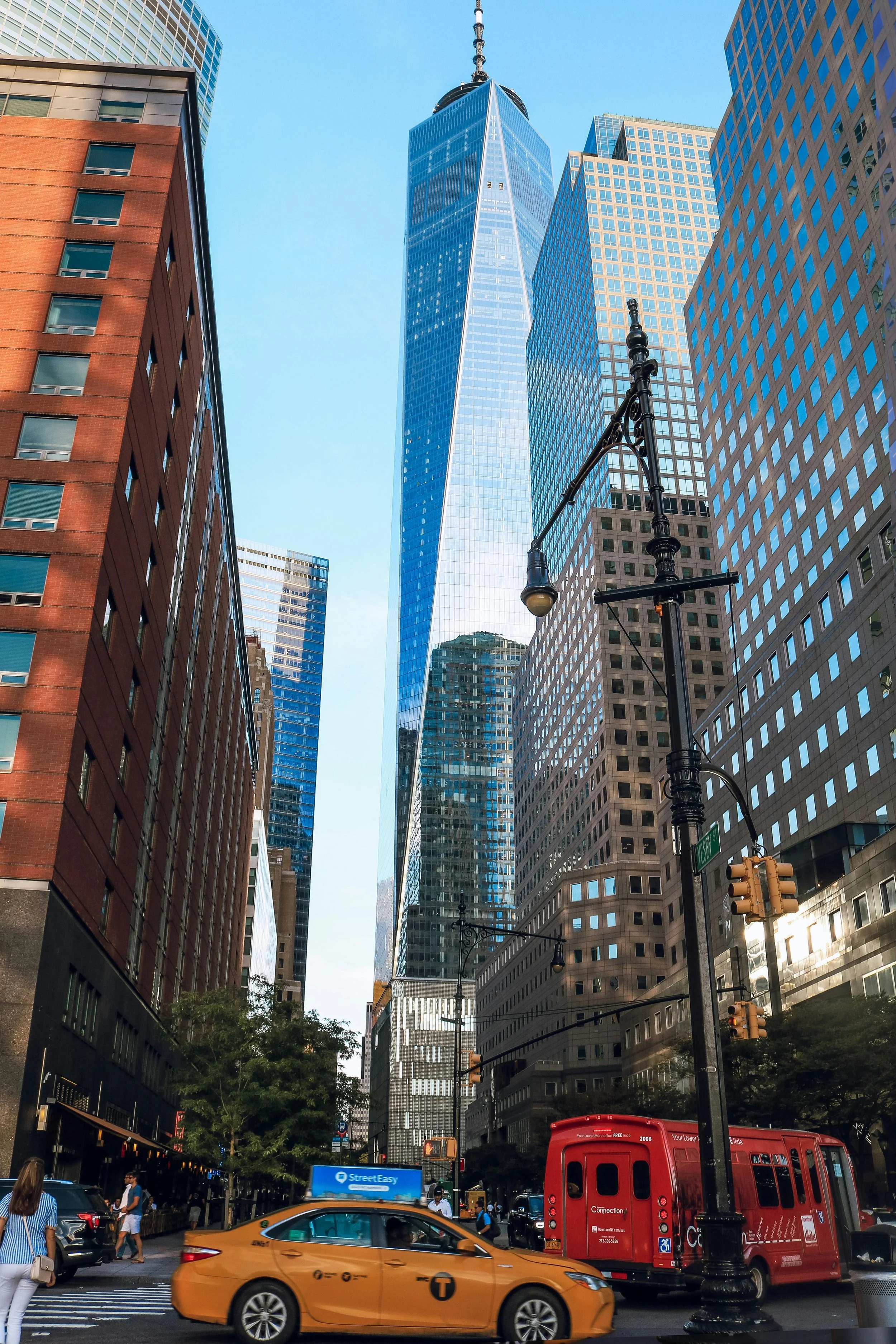 A city street scene featuring skyscrapers, including One World Trade Center, with a yellow taxi, a red bus, and pedestrians at crosswalks.