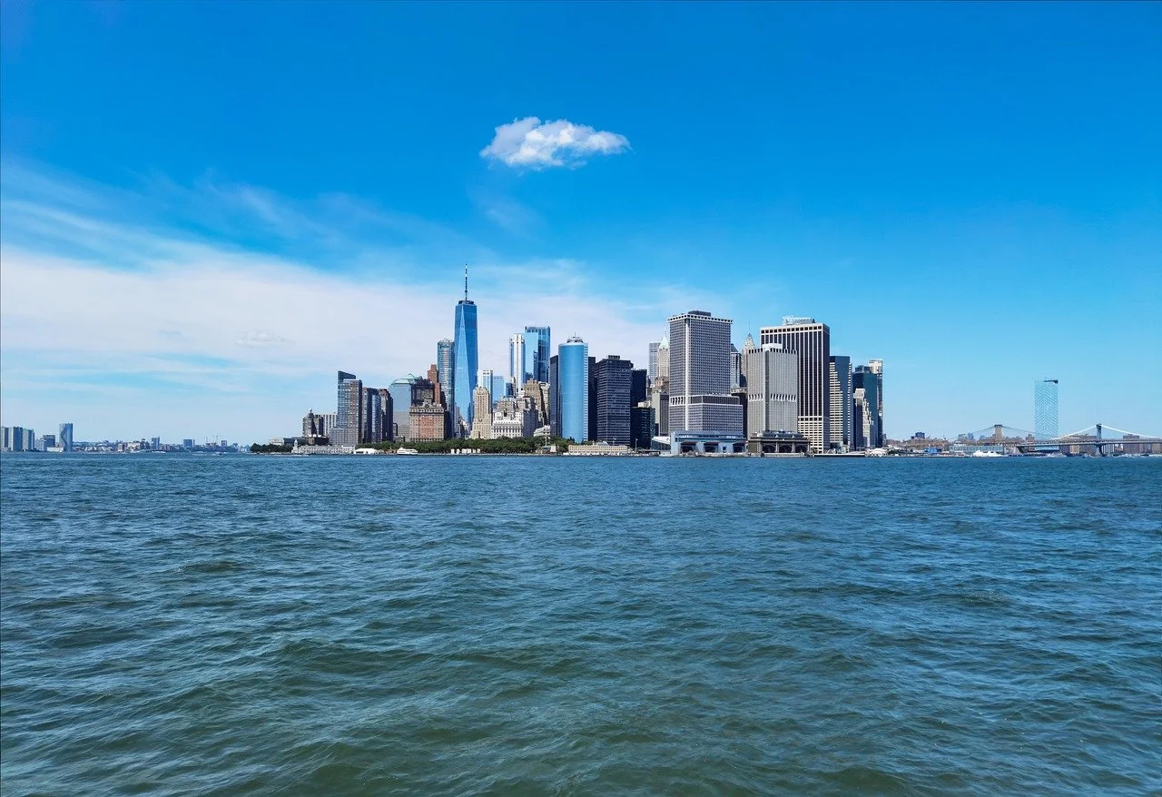 View of Manhattan skyline from across the water, blue sky with some clouds, One World Trade Center prominent in the skyline, Brooklyn Bridge visible on the right.