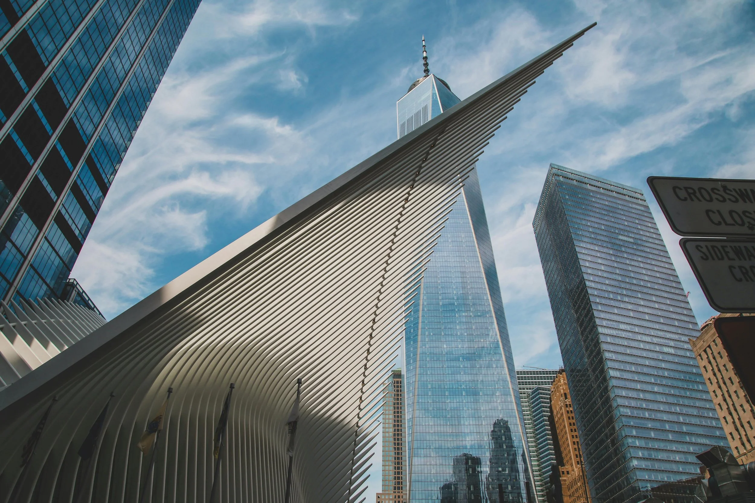Low-angle view of modern skyscrapers in New York City, including One World Trade Center and the Oculus transportation hub, against a partly cloudy sky.