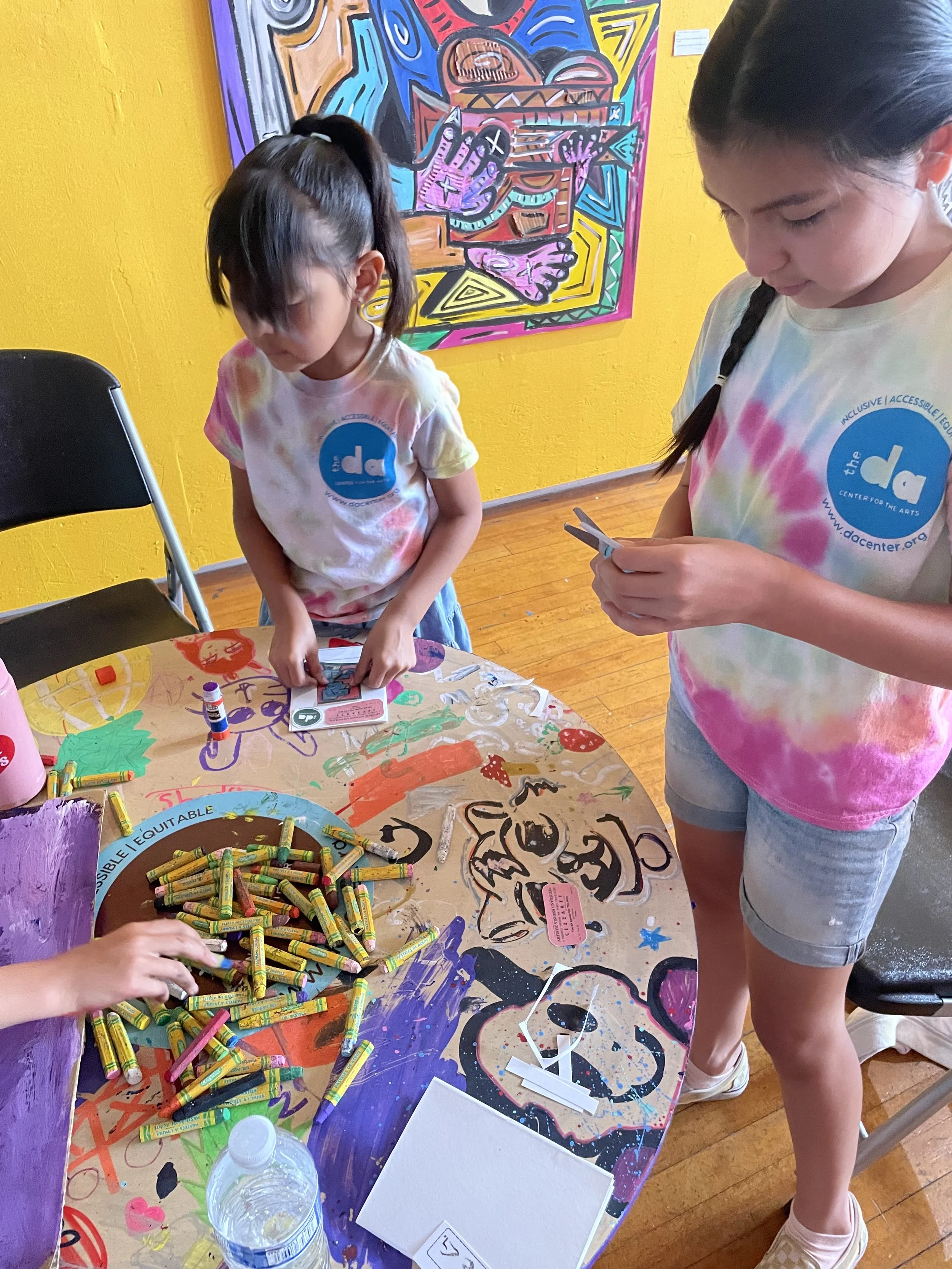 Two young girls participating in an art activity at a table covered with colorful markers, glue sticks, and paper. The table has painted and drawn designs, including a face and abstract shapes. One girl with dark hair tied in a ponytail is looking down at her project, while the other with braided hair is focused on her phone. The background features a bright yellow wall with a vibrant abstract mural.