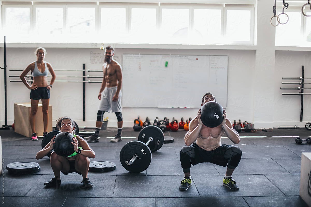 Two men doing squats with kettlebells in a gym, women and a man standing in the background, gym equipment including weights and rings visible in the background.