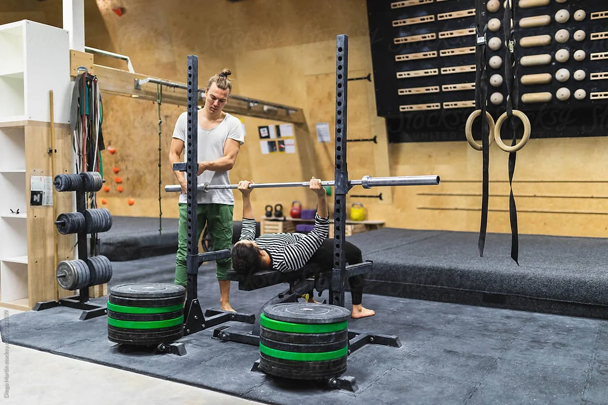 A woman performs a bench press exercise with a barbell in a gym while a spotter assists her. The gym has weights, kettlebells, and gymnastic rings.