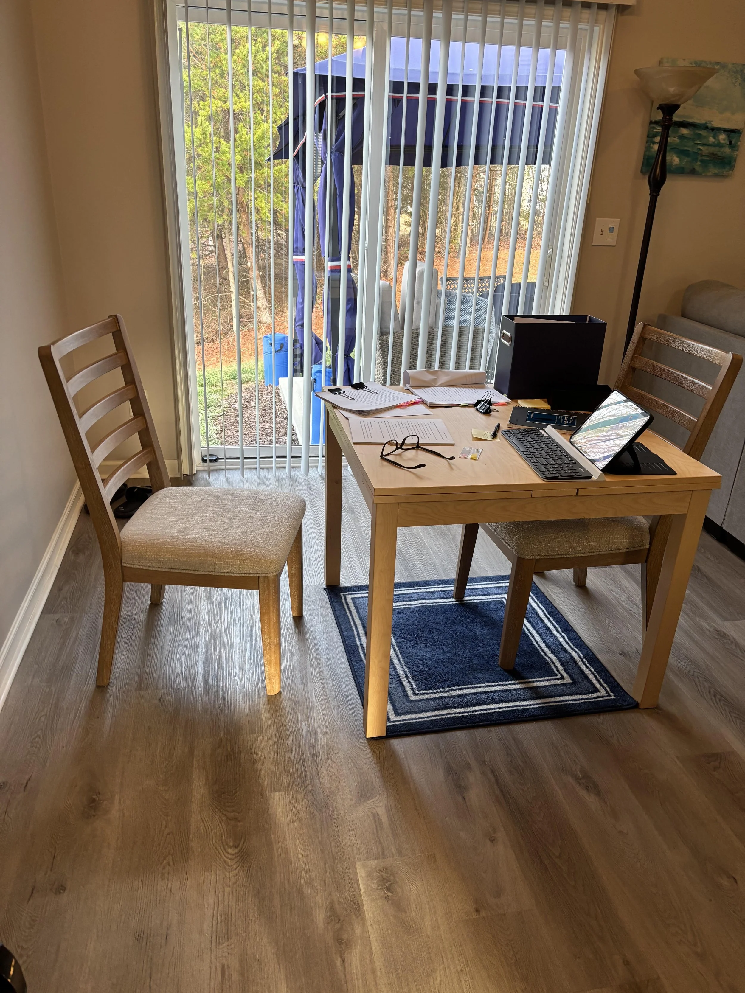 Home office with a wooden table, two chairs, a desk organizer, books, glasses, and electronic devices, with a sliding glass door leading to an outdoor patio area with a blue umbrella and trees in the background.