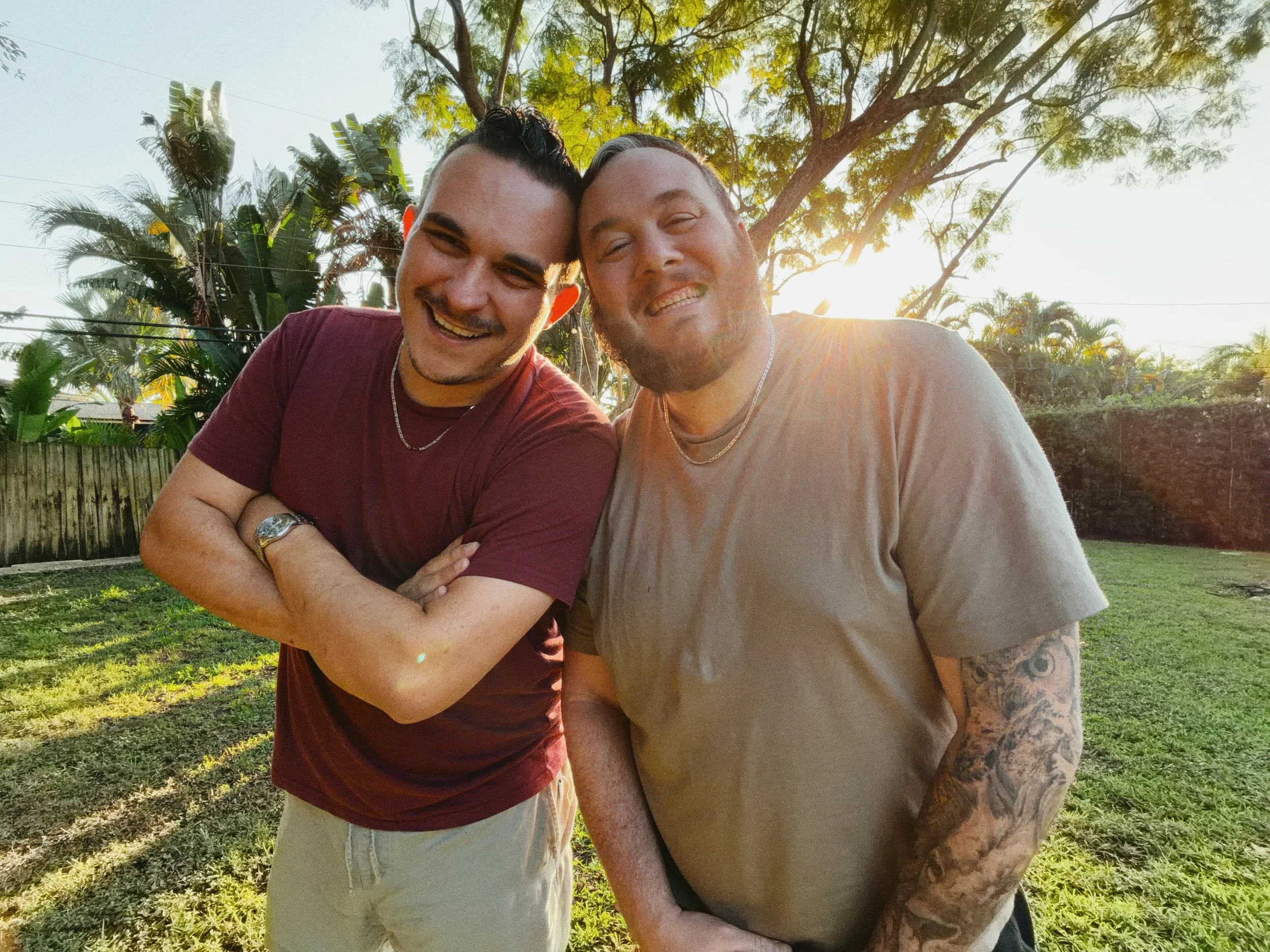 Two smiling men stand outdoors in a backyard with green grass, trees, and a wooden fence. It is sunny and the sun is setting, creating a warm glow behind them selling the best cinnamon rolls.