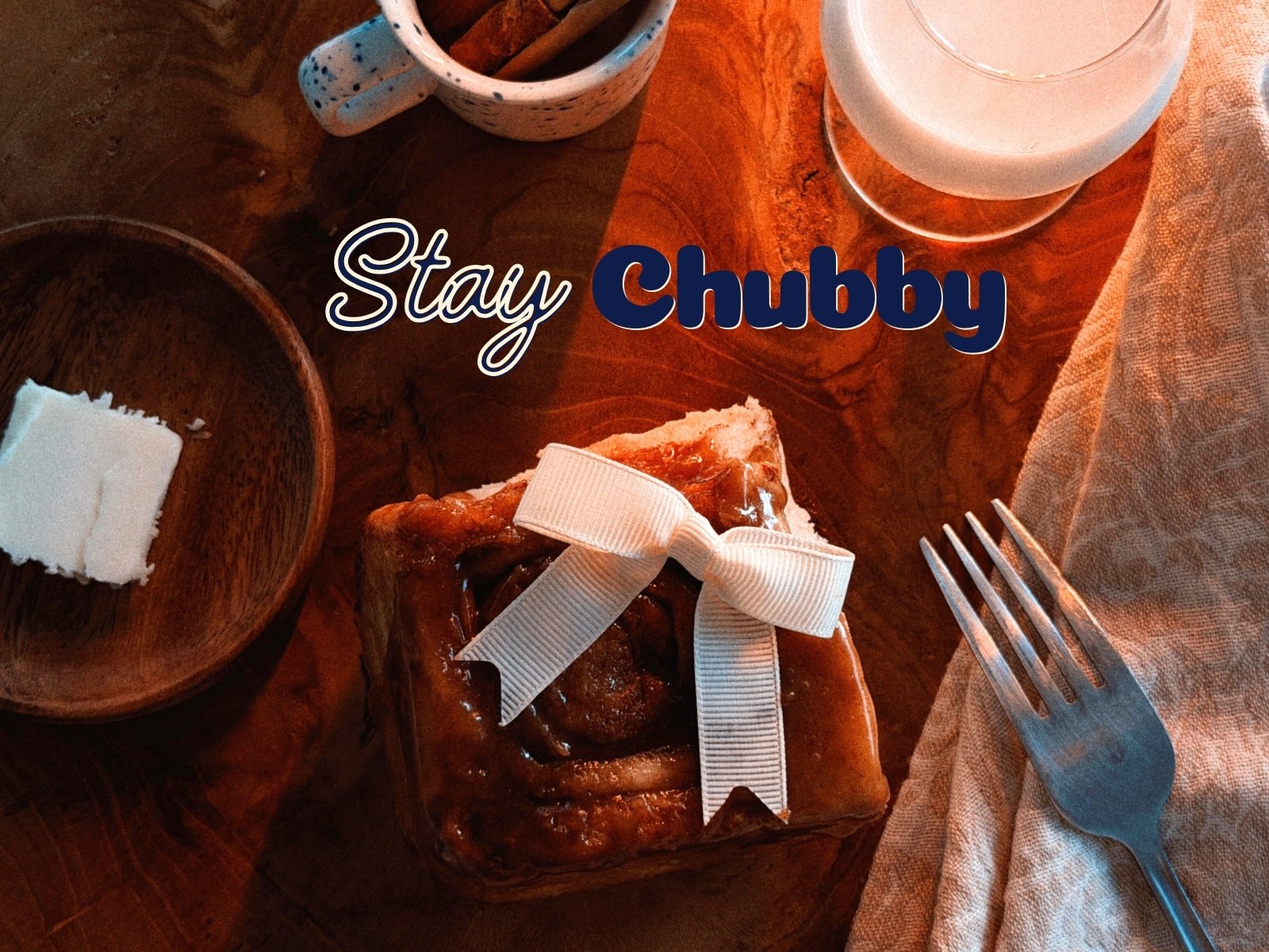 A slice of cinnamon roll with icing and a white ribbon on top, a fork, a glass of milk, a small bowl of butter, and a cup with cinnamon sticks on a wooden table with the text "Stay Chubby"